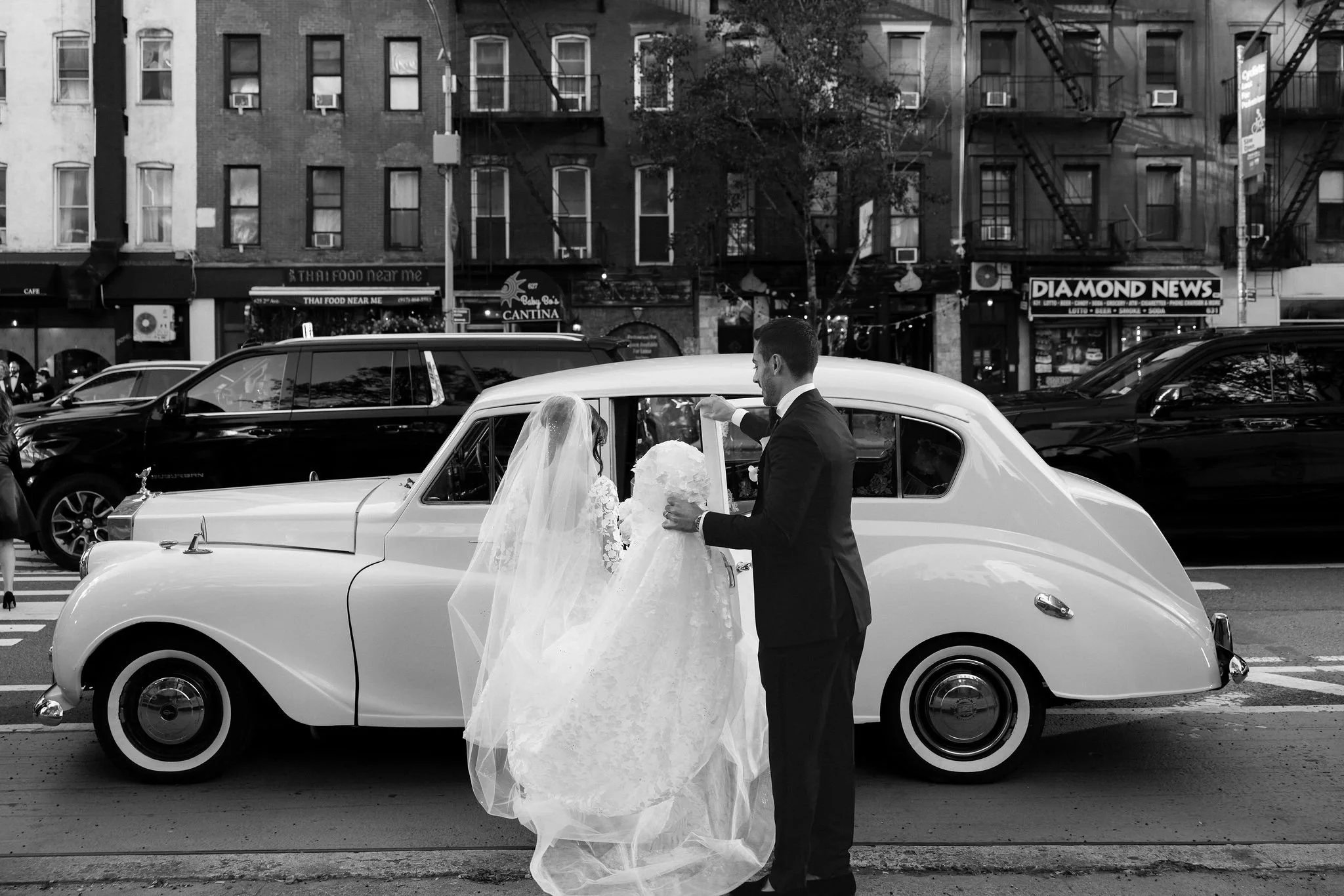 A black and white photo of a man in a suit and a woman in a wedding dress standing outside a vintage white car, with a city street full of parked and moving cars behind them. The background shows buildings with storefronts and fire escapes.