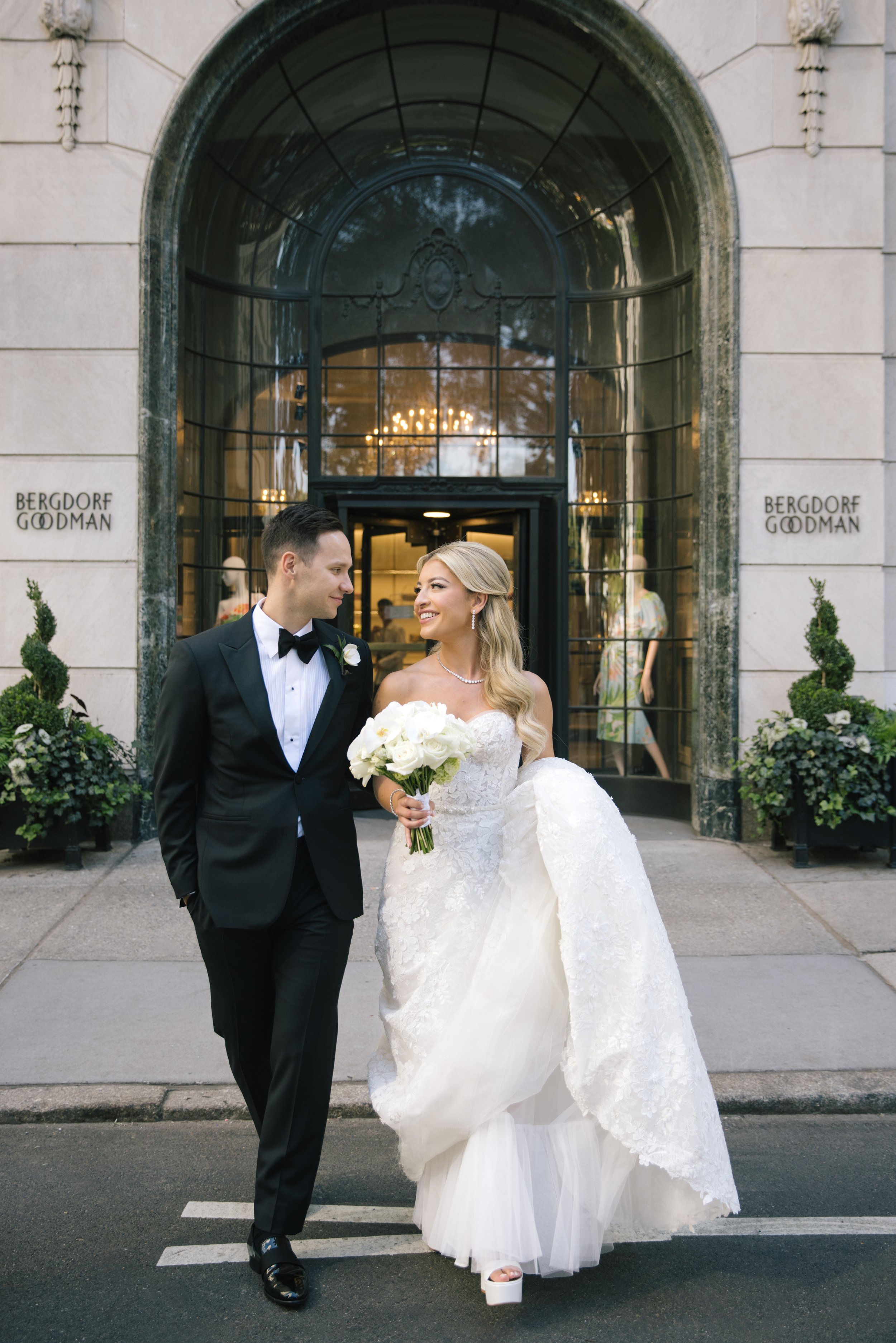 A bride and groom walking arm in arm outside a building with large glass doors and an arched window. The bride is smiling and holding a bouquet of white roses, wearing a strapless lace wedding gown with a tulle skirt and platforms. The groom is dressed in a black tuxedo with a white shirt and bow tie, looking at the bride. The building has beige stone walls with decorative stonework and signs reading