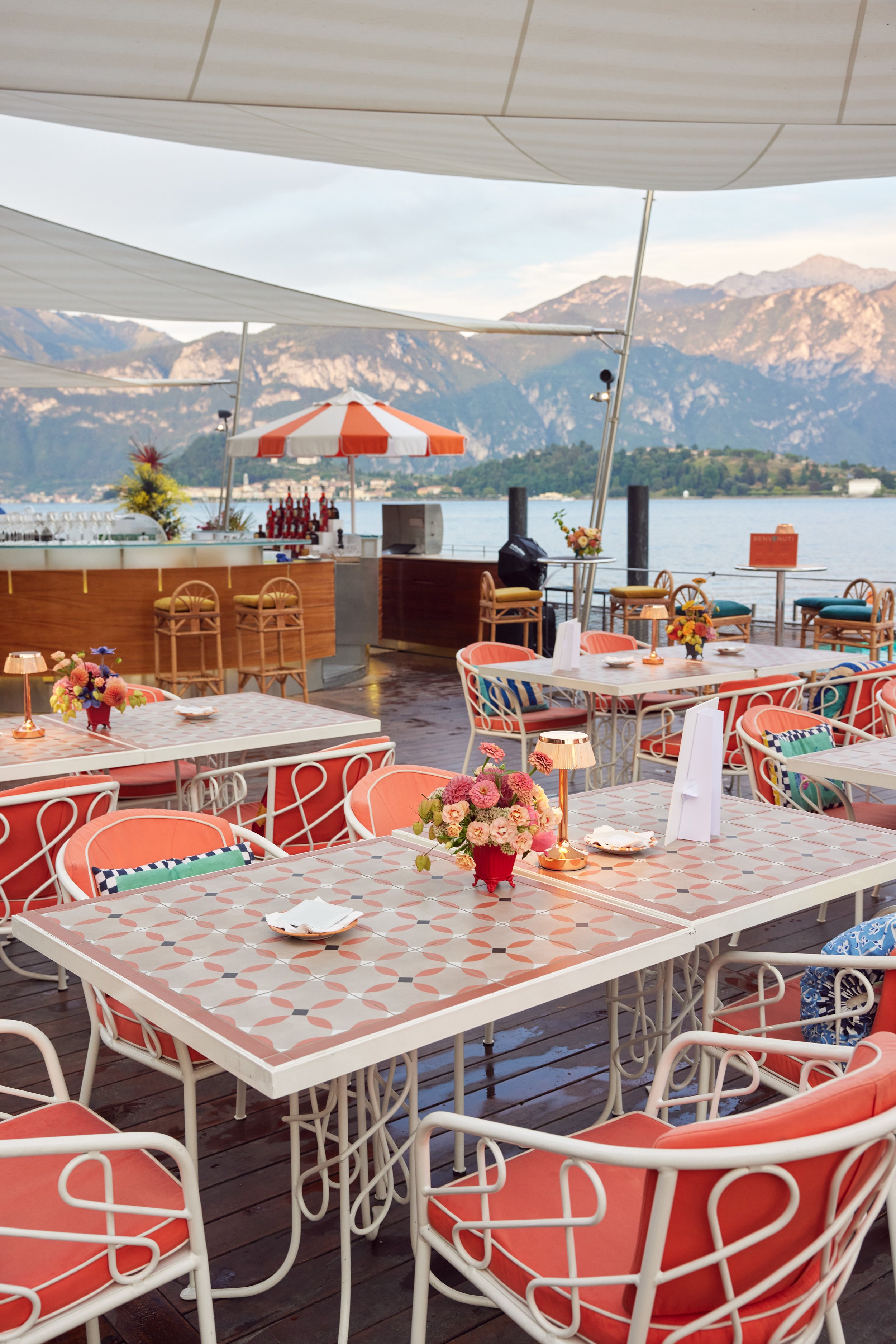 Outdoor restaurant or cafe with tables and chairs, decorated with flowers, overlooking a body of water with mountains in the background, under white and orange striped umbrellas.