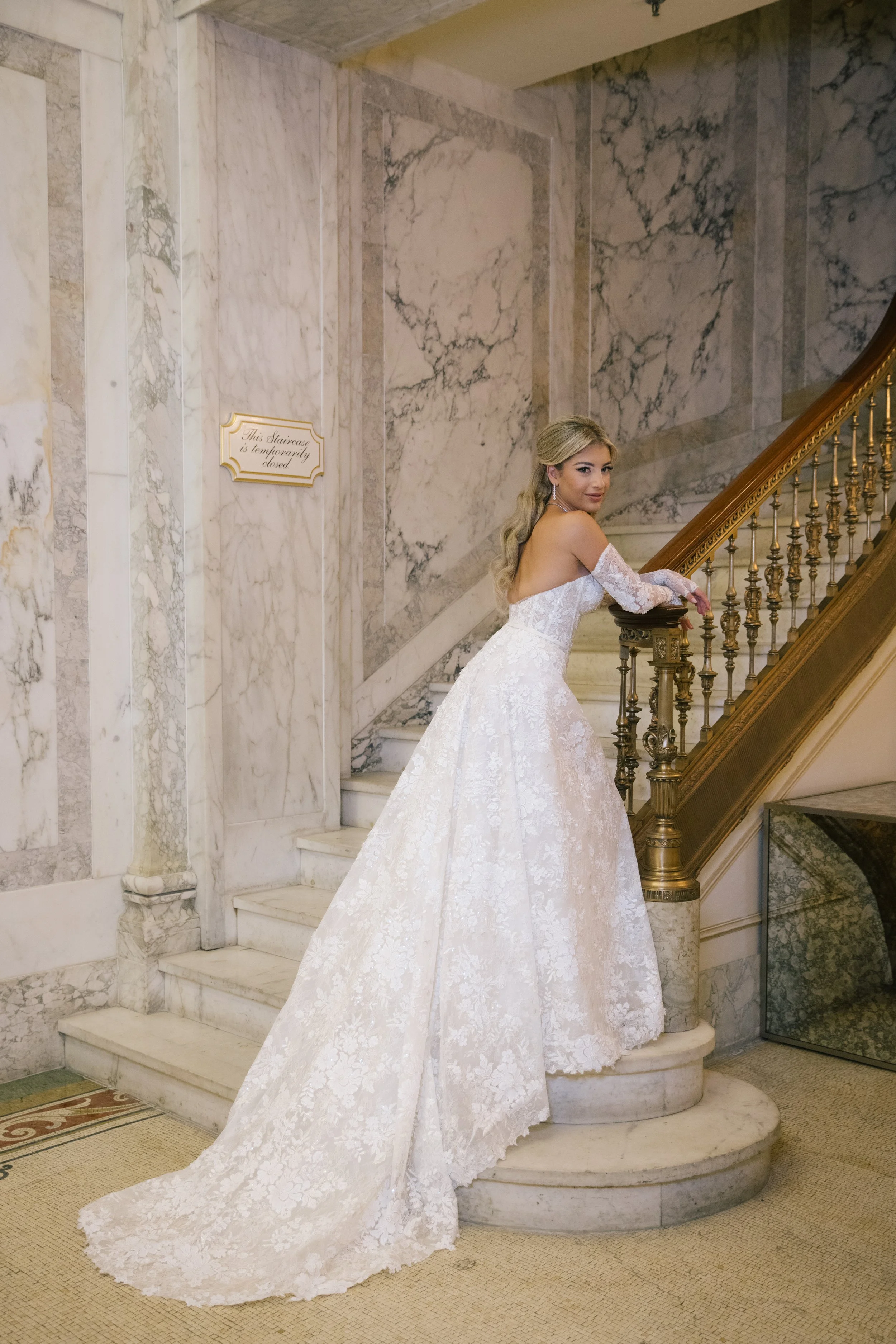 A woman in a white lace wedding gown leaning on a decorative staircase rail inside a marble-lined building.