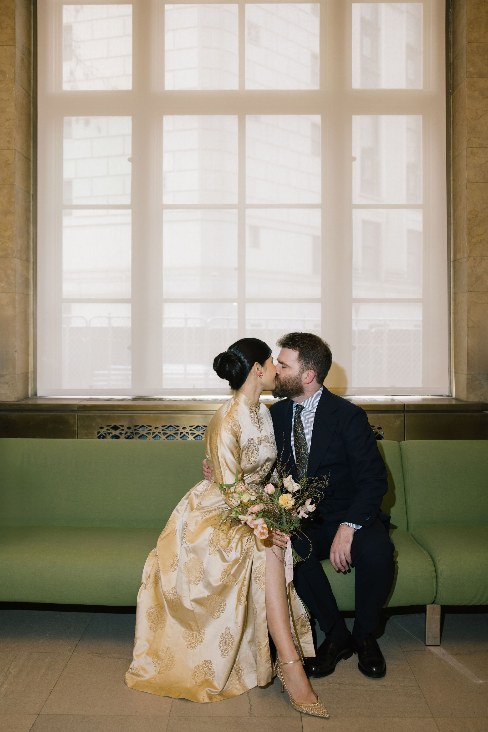 A man and woman, dressed in formal attire, share a kiss while seated on a green bench with a large window behind them. The woman wears a gold gown and high heels, holding a bouquet of flowers, and the man wears a dark suit and tie. The window is framed by beige stone walls.