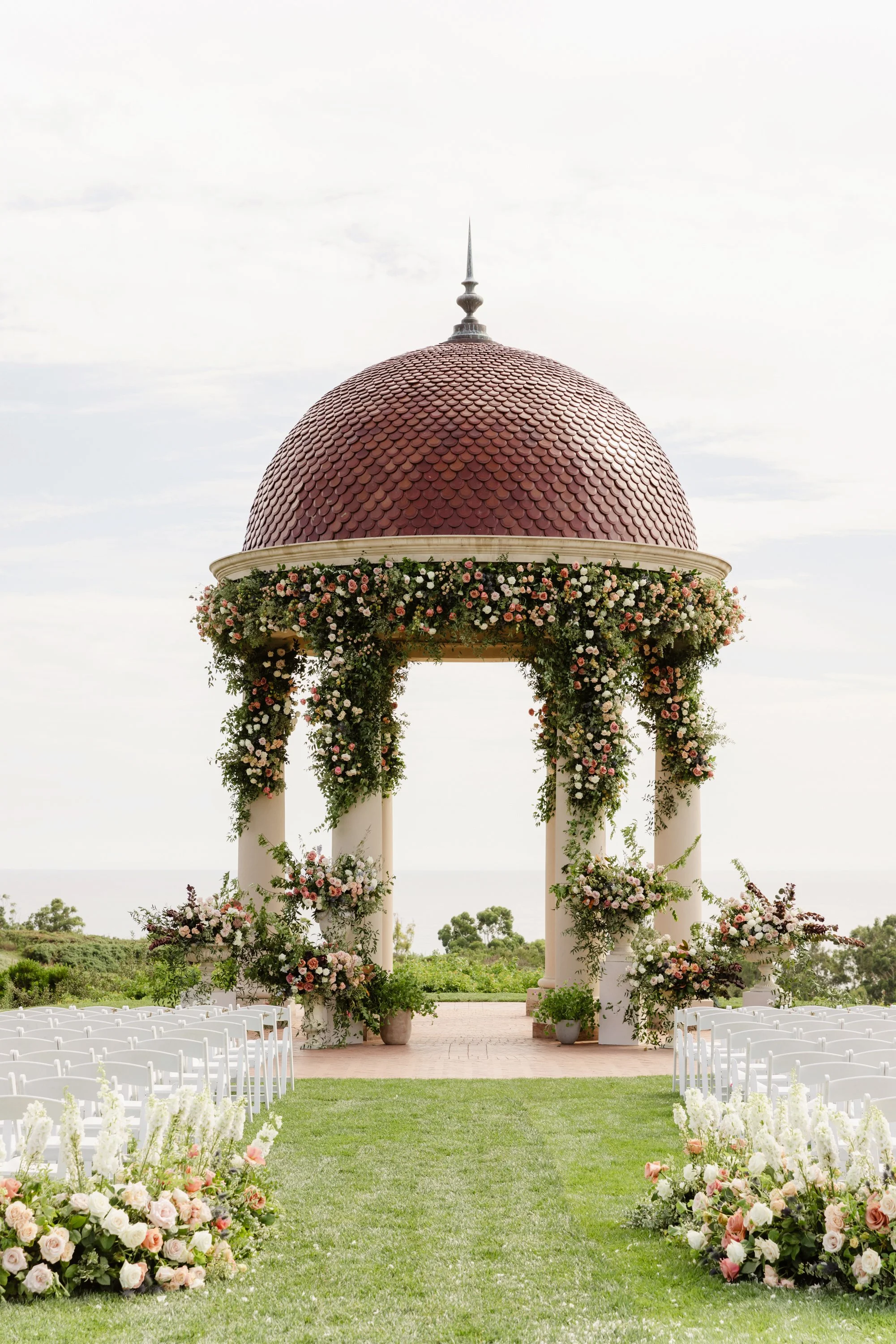 A wedding ceremony setup outdoors with floral arrangements and a domed structure with a spire, on a grassy area with chairs.