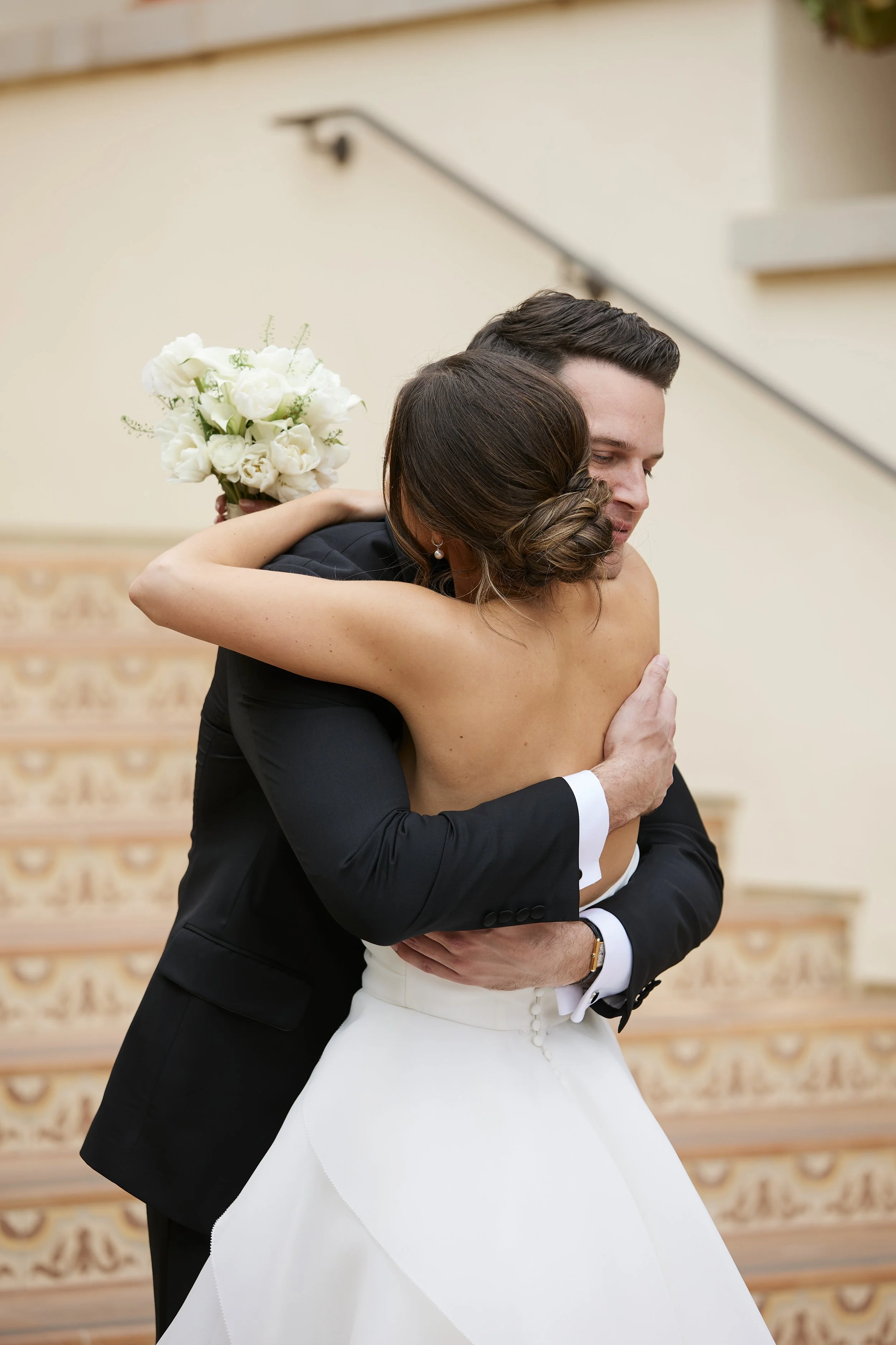 A couple, dressed in wedding attire, embracing each other. The woman is wearing a white wedding gown with a draped skirt, and the man is wearing a black tuxedo with white shirt and black bow tie. The woman is holding a bouquet of white flowers behind