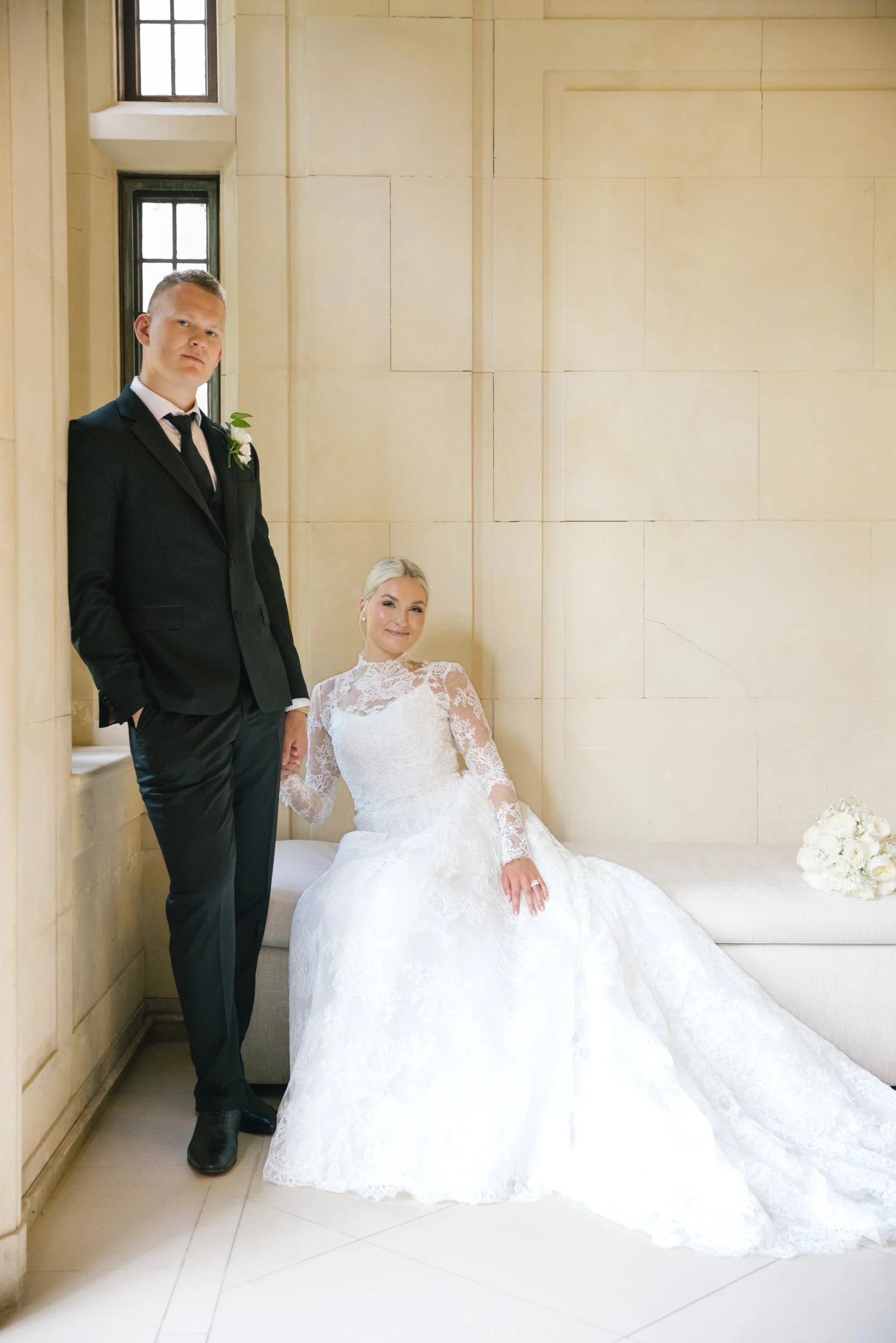 A groom in a black suit and tie standing next to a bride in a white lace wedding gown, who is sitting on a bench against a beige wall with windows.