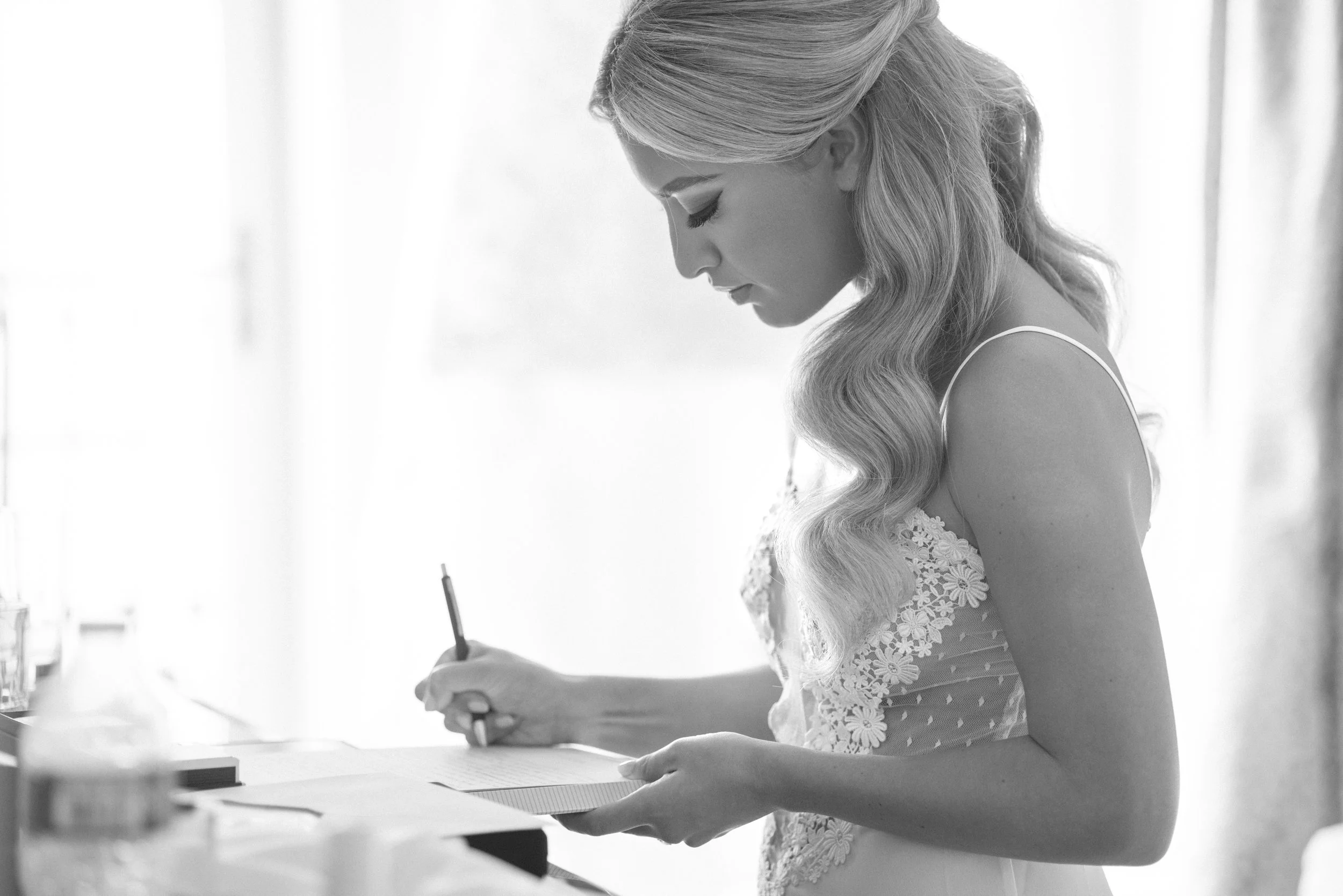 A woman with long wavy hair wearing a lace dress, sitting at a table, writing on paper with a pen in a bright room.