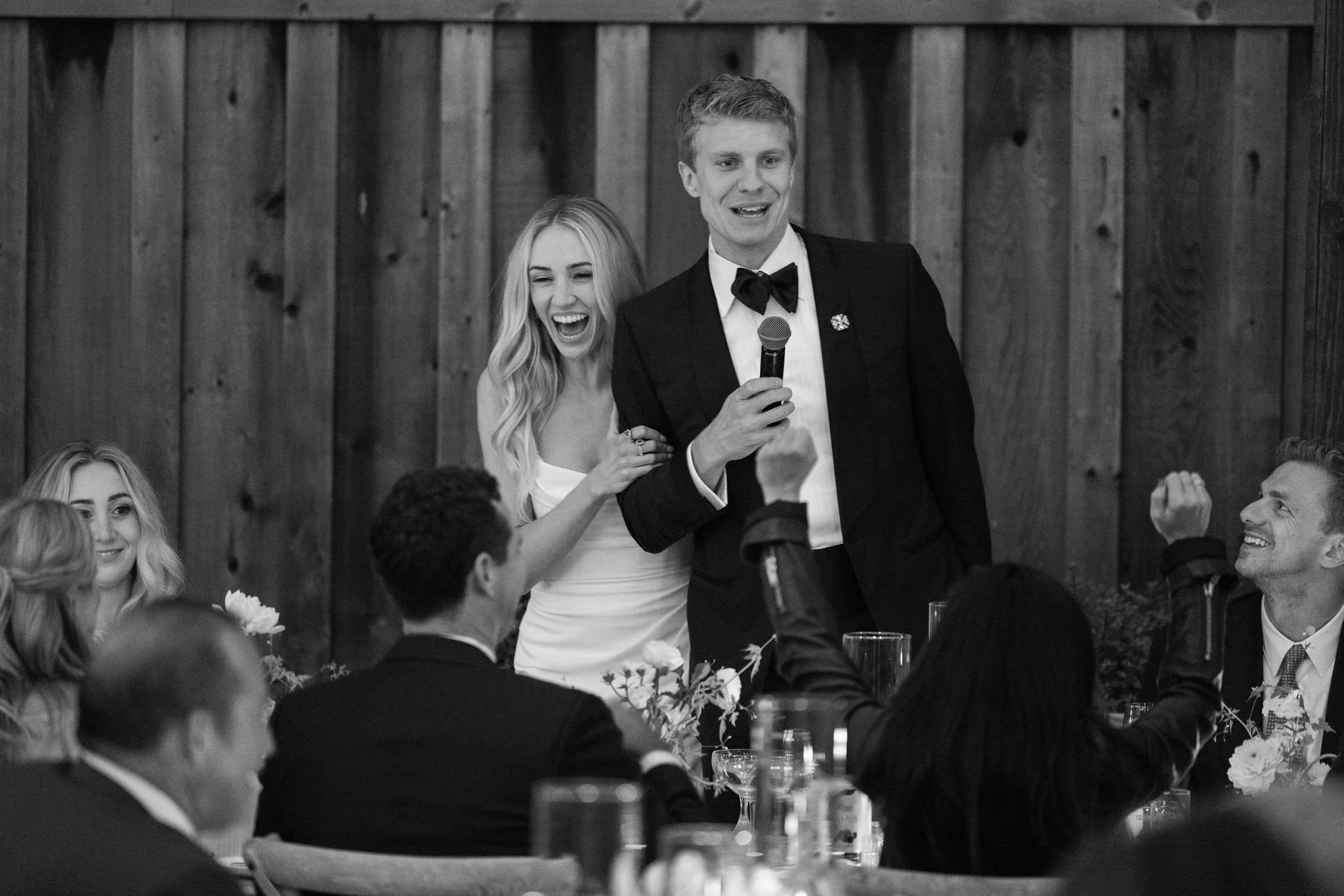 A man in a tuxedo with a bowtie is speaking into a microphone at a formal event. A woman standing next to him is smiling and appears to be laughing. Other guests are seated at tables with flowers and glasses, listening and smiling. The background fea