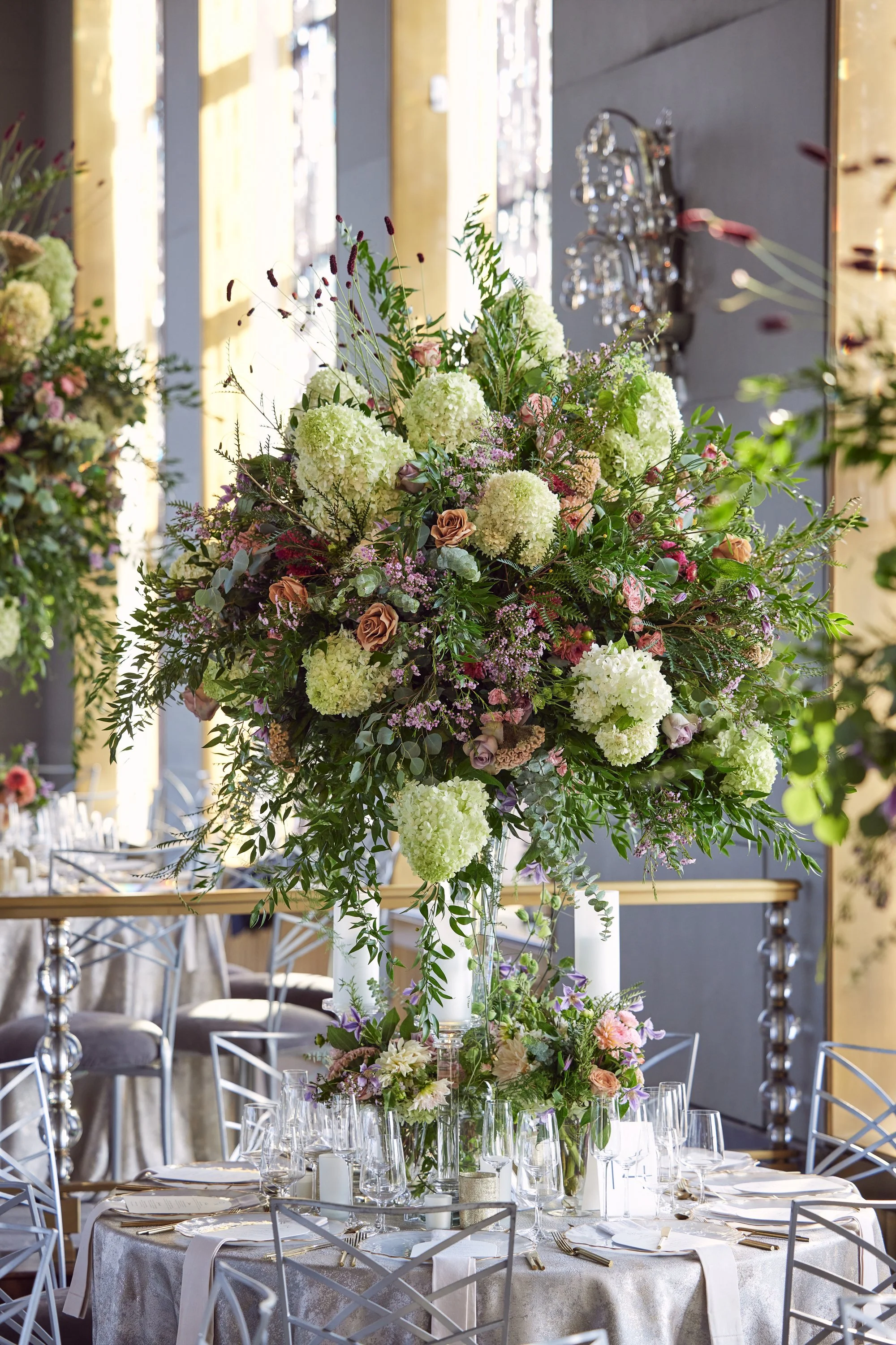 Elegant indoor wedding or event reception with a large floral centerpiece consisting of white hydrangeas, roses, and greenery, with tables set with white tablecloths, glasses, and silverware. Metallic chairs are arranged around the tables, and natura