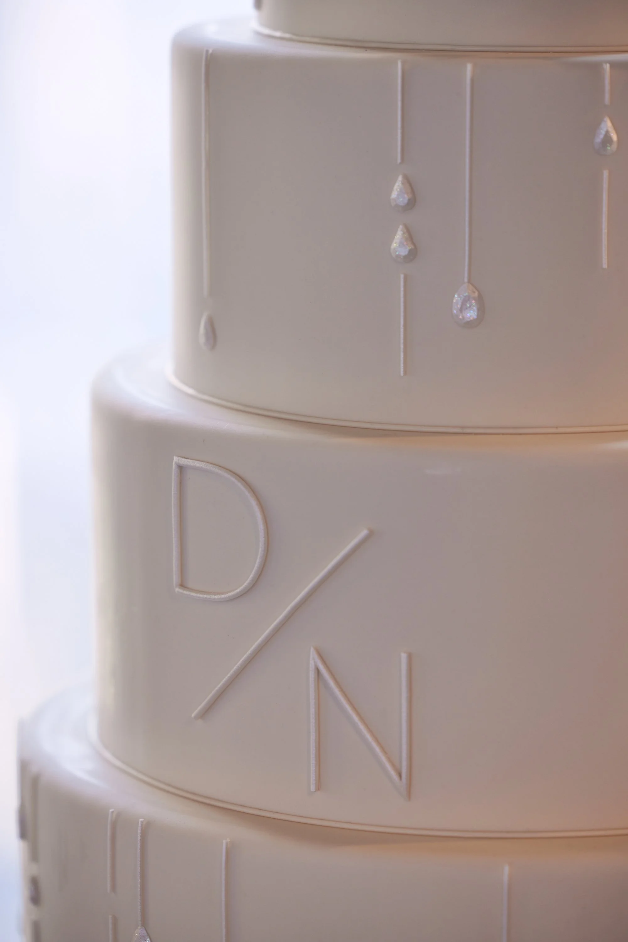 Close-up of a white tiered wedding cake with embroidered initials and dangling gemstone decorations.