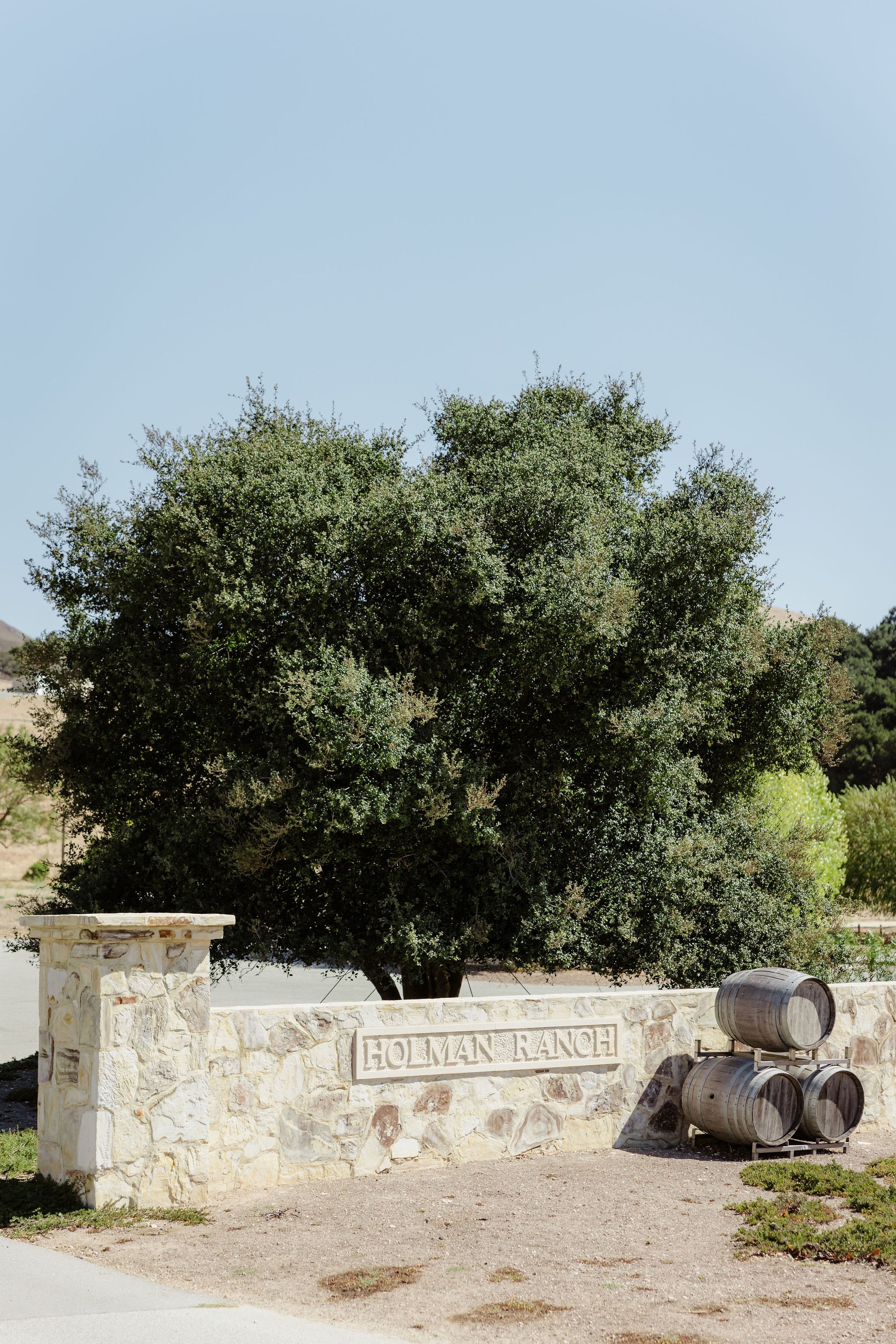 Stone entrance sign that reads 'Holman Ranch,' with three stacked barrels on a metal stand nearby, large tree behind the sign, and a clear blue sky above.