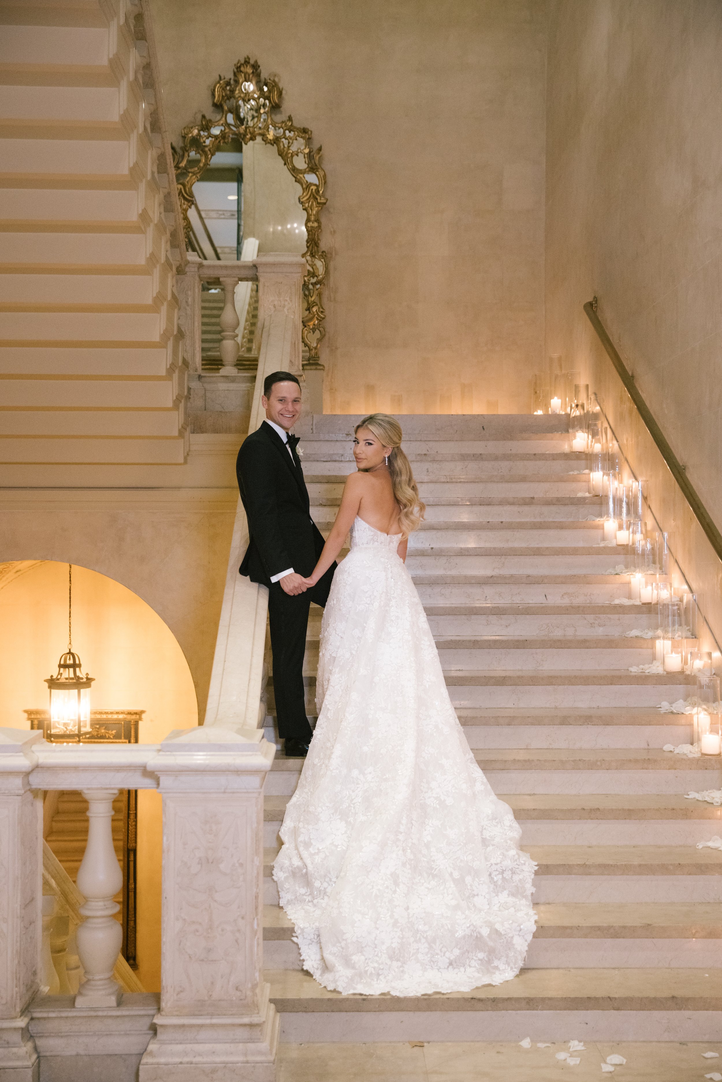 A newlywed couple holding hands and smiling on a staircase in a elegant, historic building. The groom is wearing a black tuxedo and the bride is in a white lace wedding gown with long blonde hair.