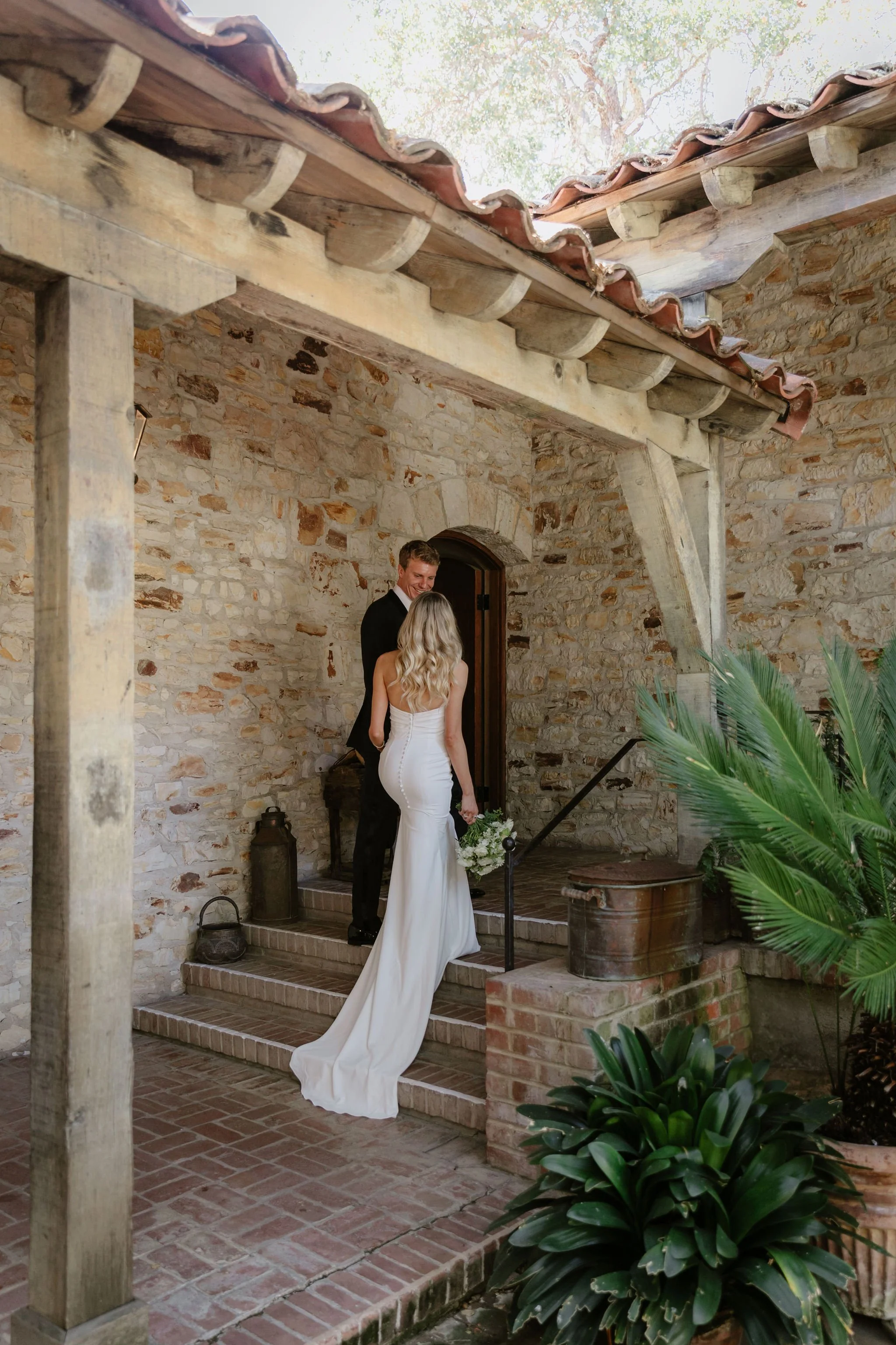 A bride and groom stand on brick steps outside a rustic stone building, holding hands and facing each other. The bride wears a white dress with a train, and the groom wears a black suit. The setting is decorated with green plants and old artifacts, c