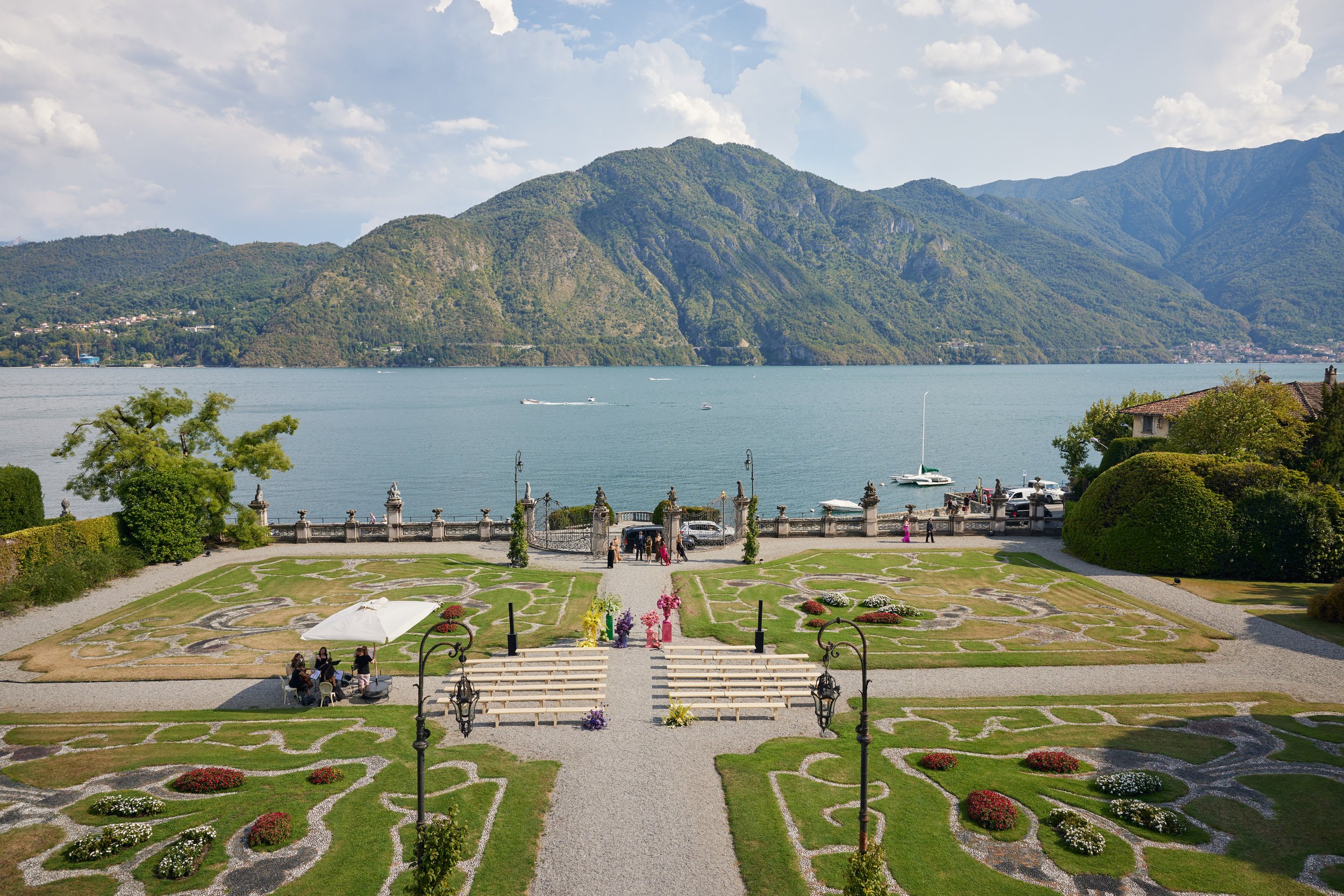 View of a landscaped garden with flower beds, benches, and a gate in the foreground, a lake with boats and hills in the background, under partly cloudy skies.