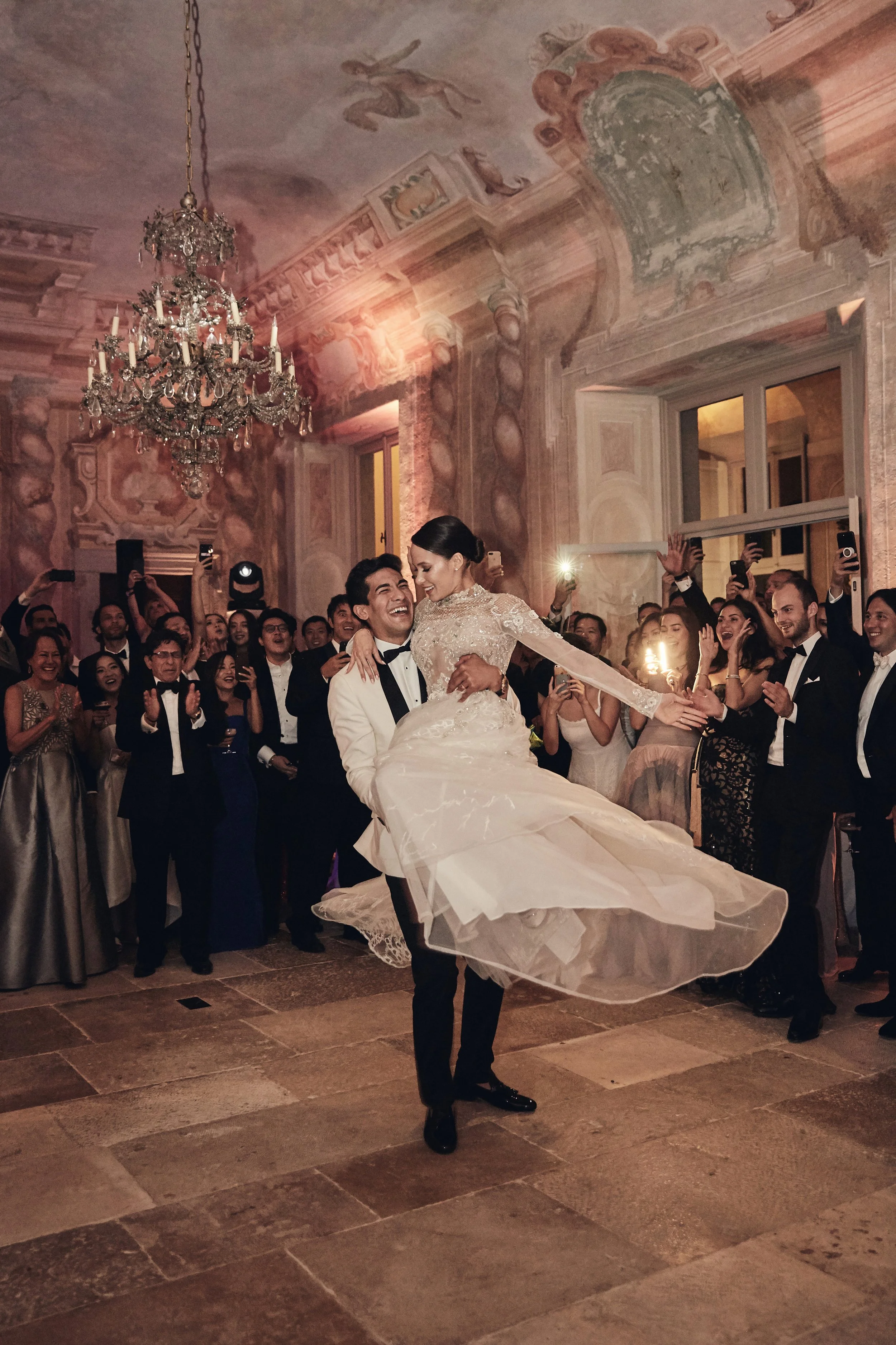 A newlywed couple dances at their wedding reception, surrounded by guests in an elegant hall with ornate decorations and a chandelier.