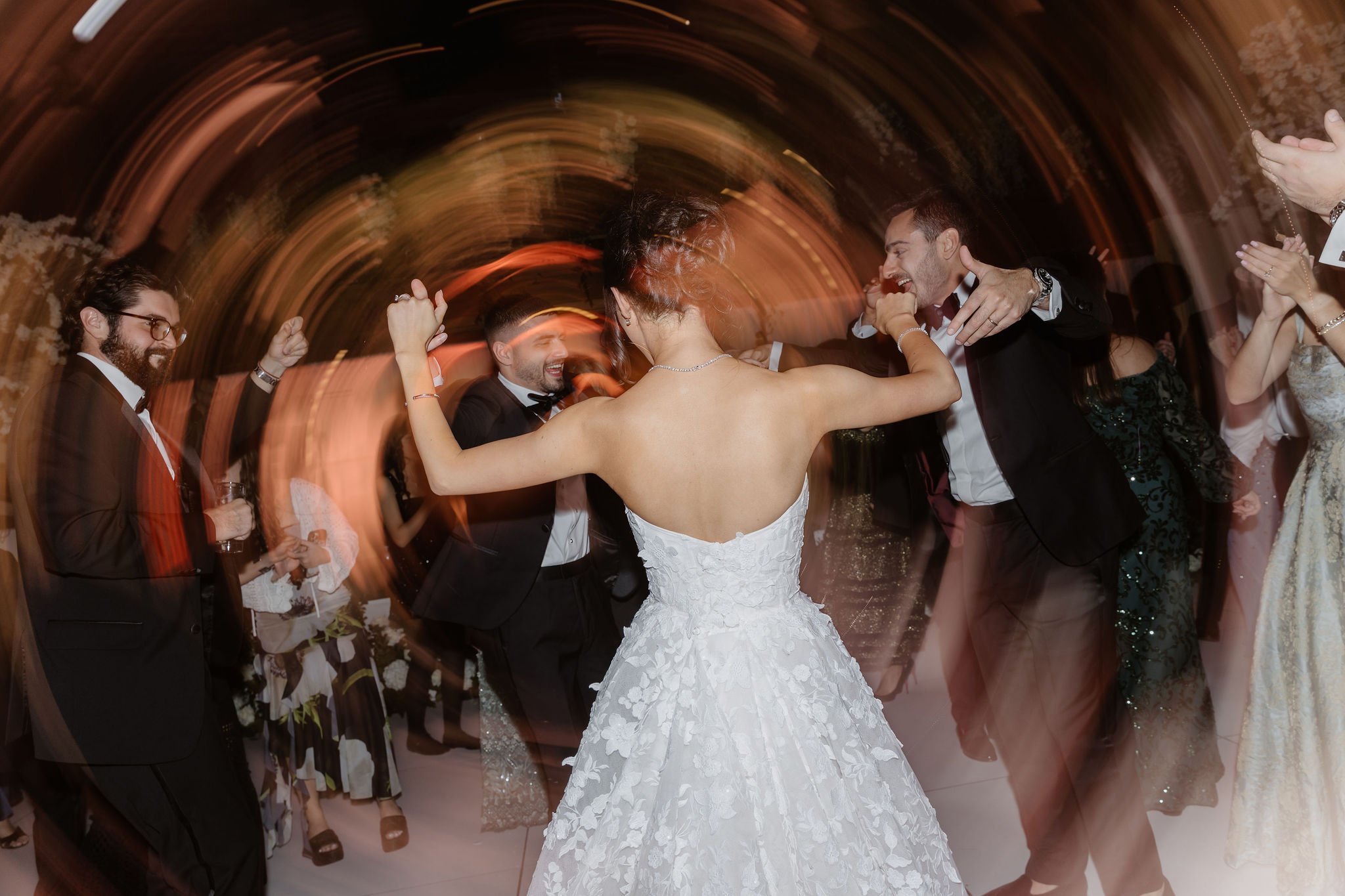 People dancing at a wedding reception, with a bride in a white gown and guests in formal attire, swirling around under warm lighting.