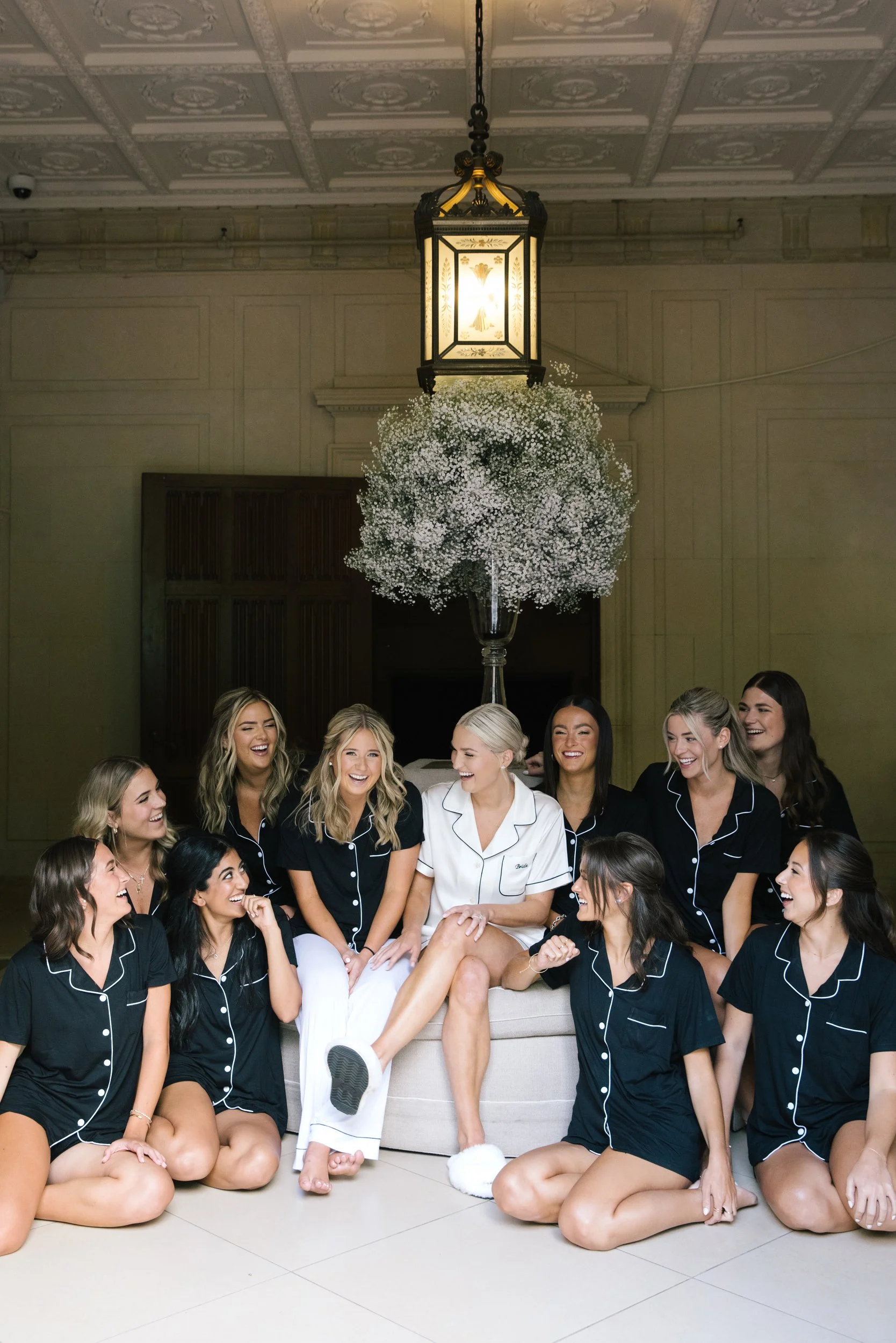 A group of women in black pajamas with white piping, one woman in white pajamas, all smiling and sitting on the floor in front of a sofa. There is a large glass vase with white baby's breath flowers hanging above them, attached to a ceiling with orna