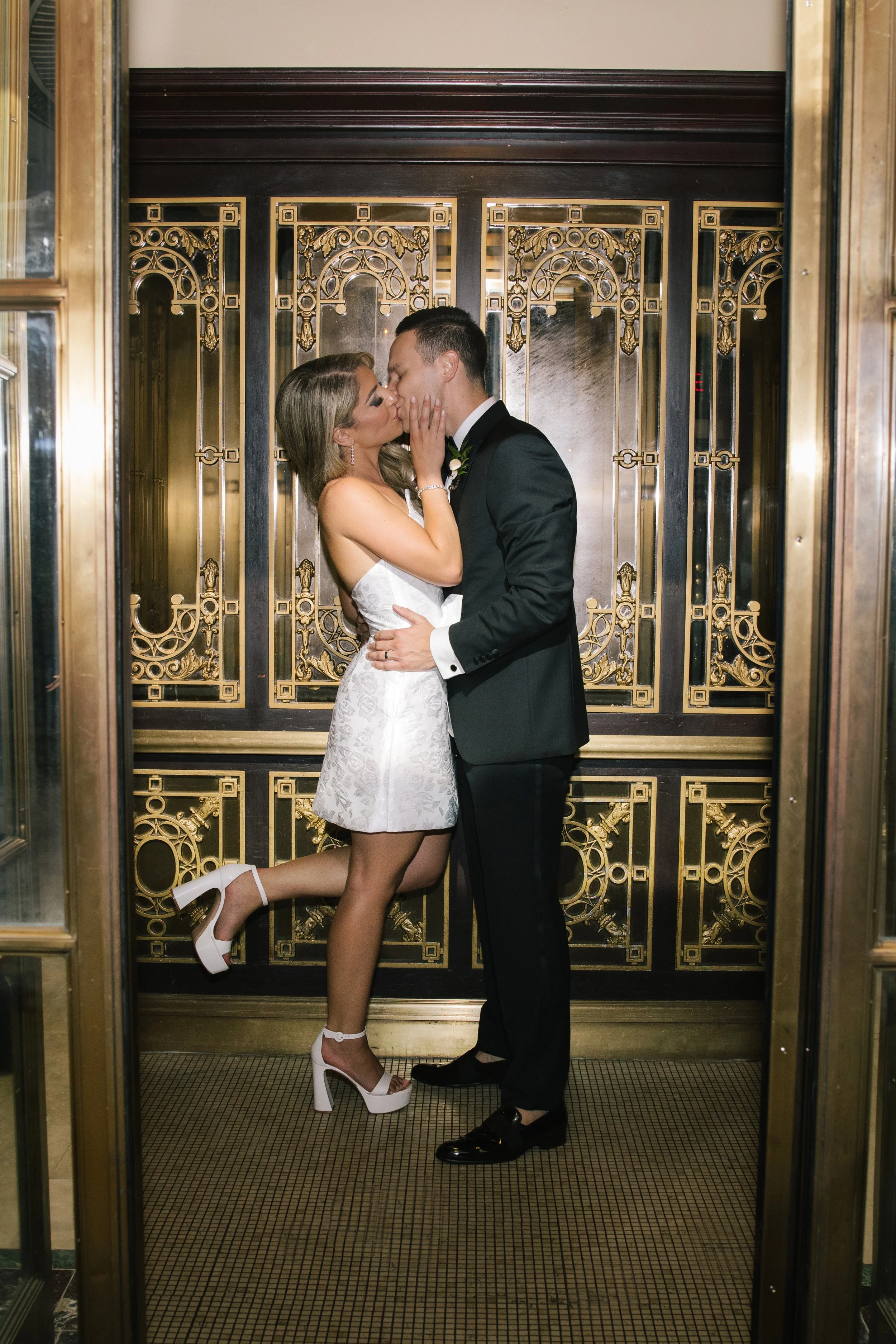A couple kissing in an elevator with ornate gold and black decor. The woman is kneeling on one leg, wearing a white dress and high heels, and the man is in a black suit and dress shoes.
