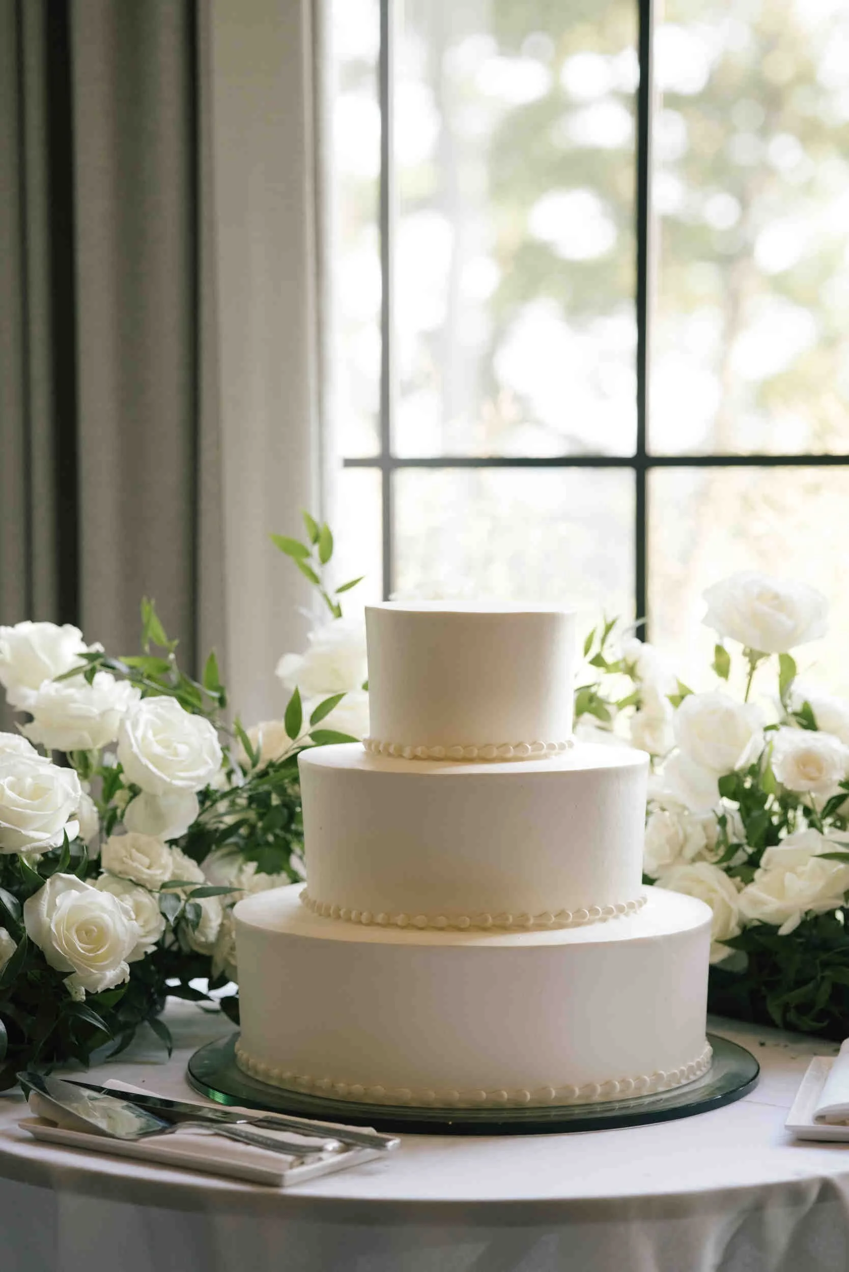 A white flower cake with three tiers, decorated with pearl-like icing, surrounded by white roses and greenery, placed on a table with silver utensils and a white tablecloth, in front of a window with a blurred outdoor scene.