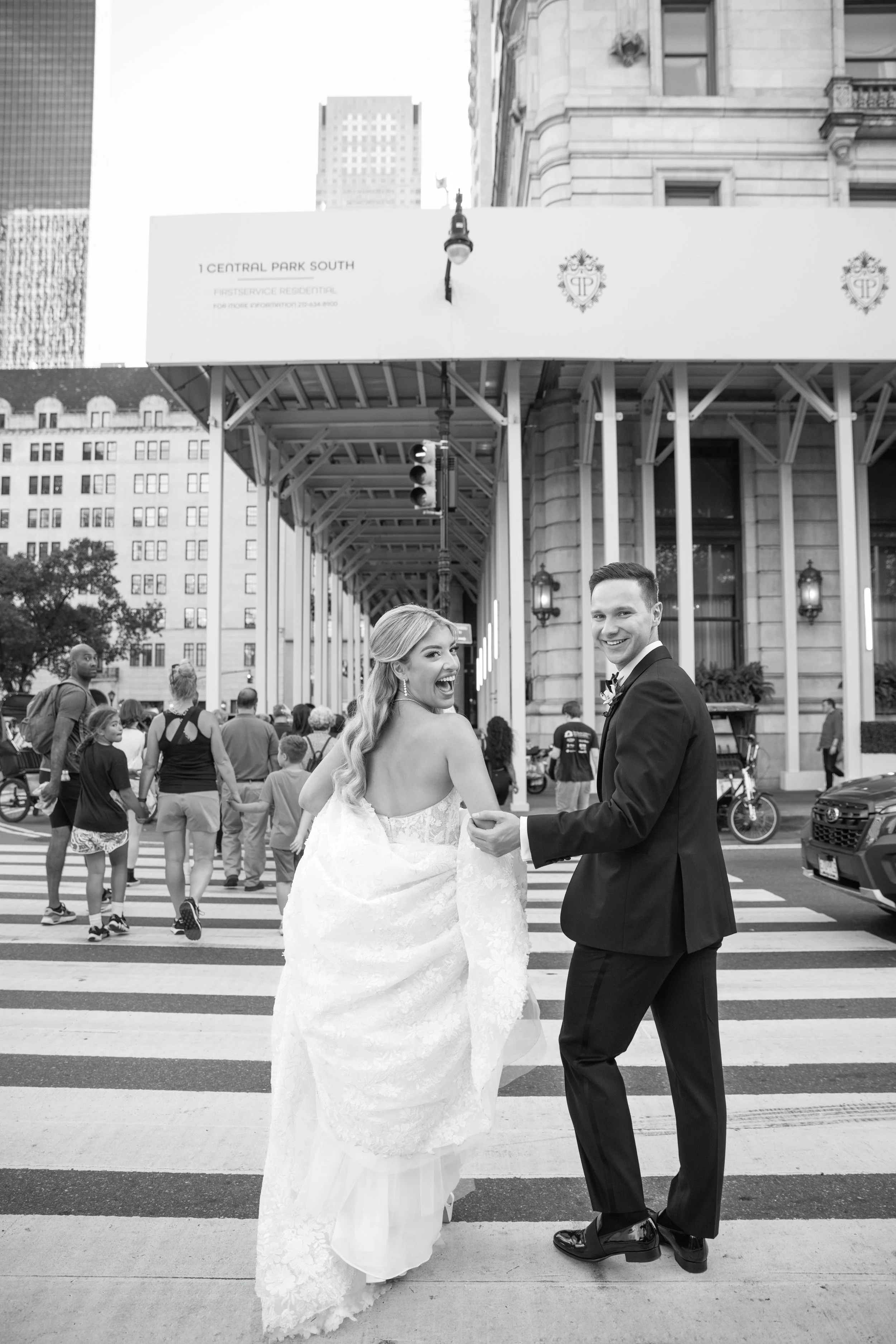 A newlywed couple holding hands and smiling at each other at a crosswalk, with a crowd of passersby and city buildings in the background, black-and-white photo.