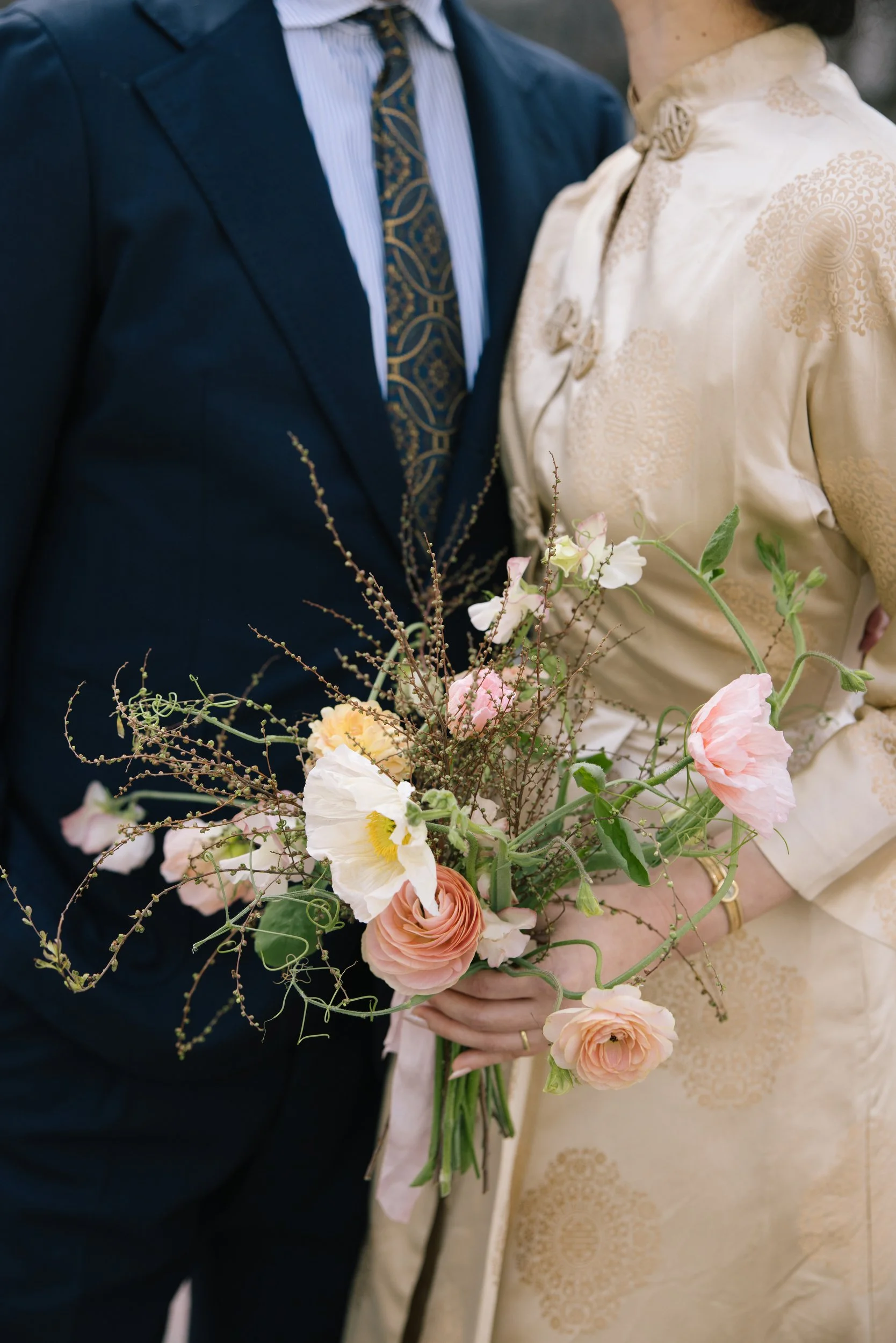 A bride and groom holding a bouquet of pink and white flowers, dressed in wedding attire, close together.