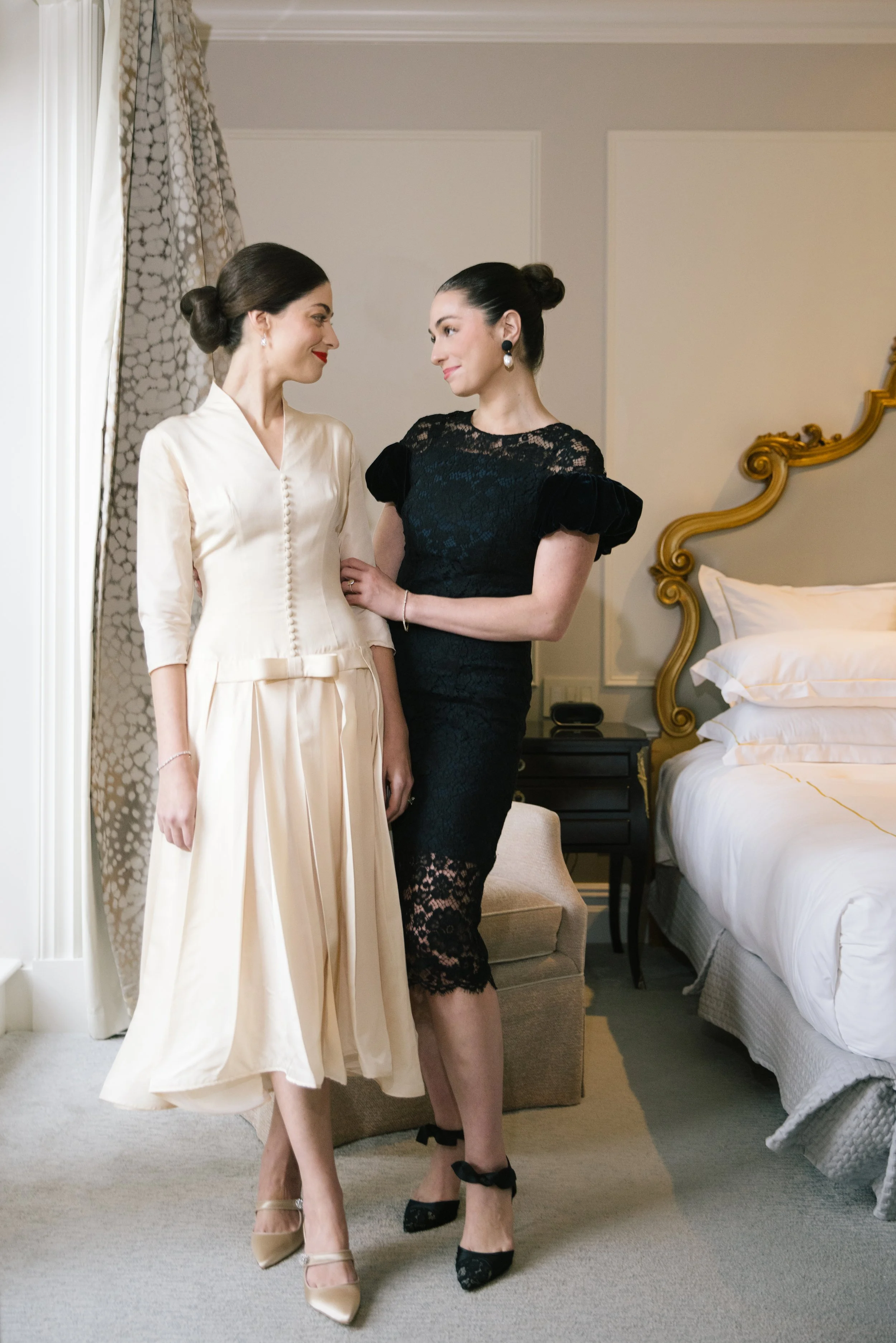Two women in elegant dresses standing close in a well-decorated bedroom, looking at each other. One is in a cream satin dress with a bow at the waist and beige heels, the other in a black lace dress with puffy sleeves and black heels with ankle bows.