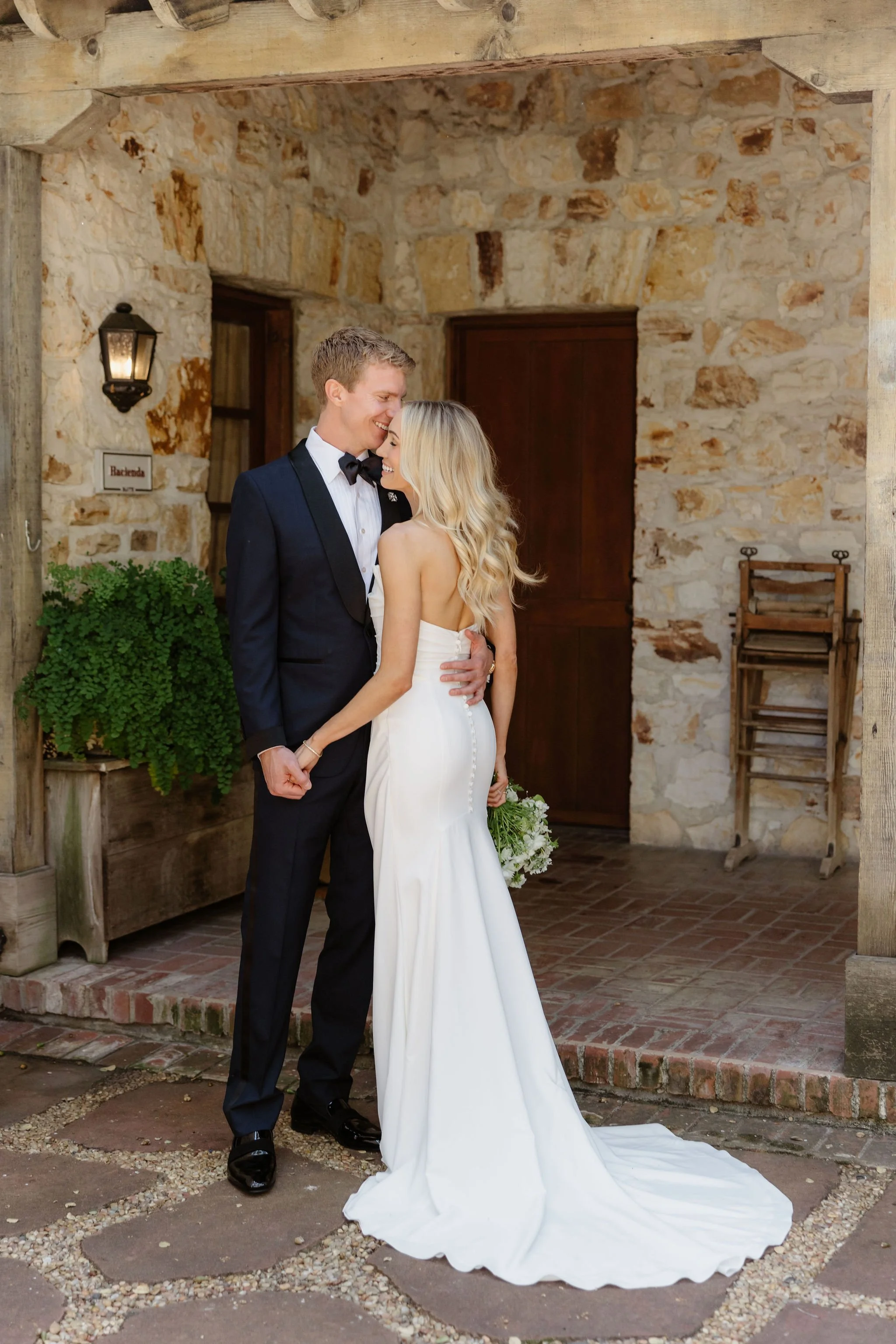 A bride and groom sharing a moment outdoors, standing on a brick patio in front of a rustic stone building, with the bride holding a bouquet of flowers.