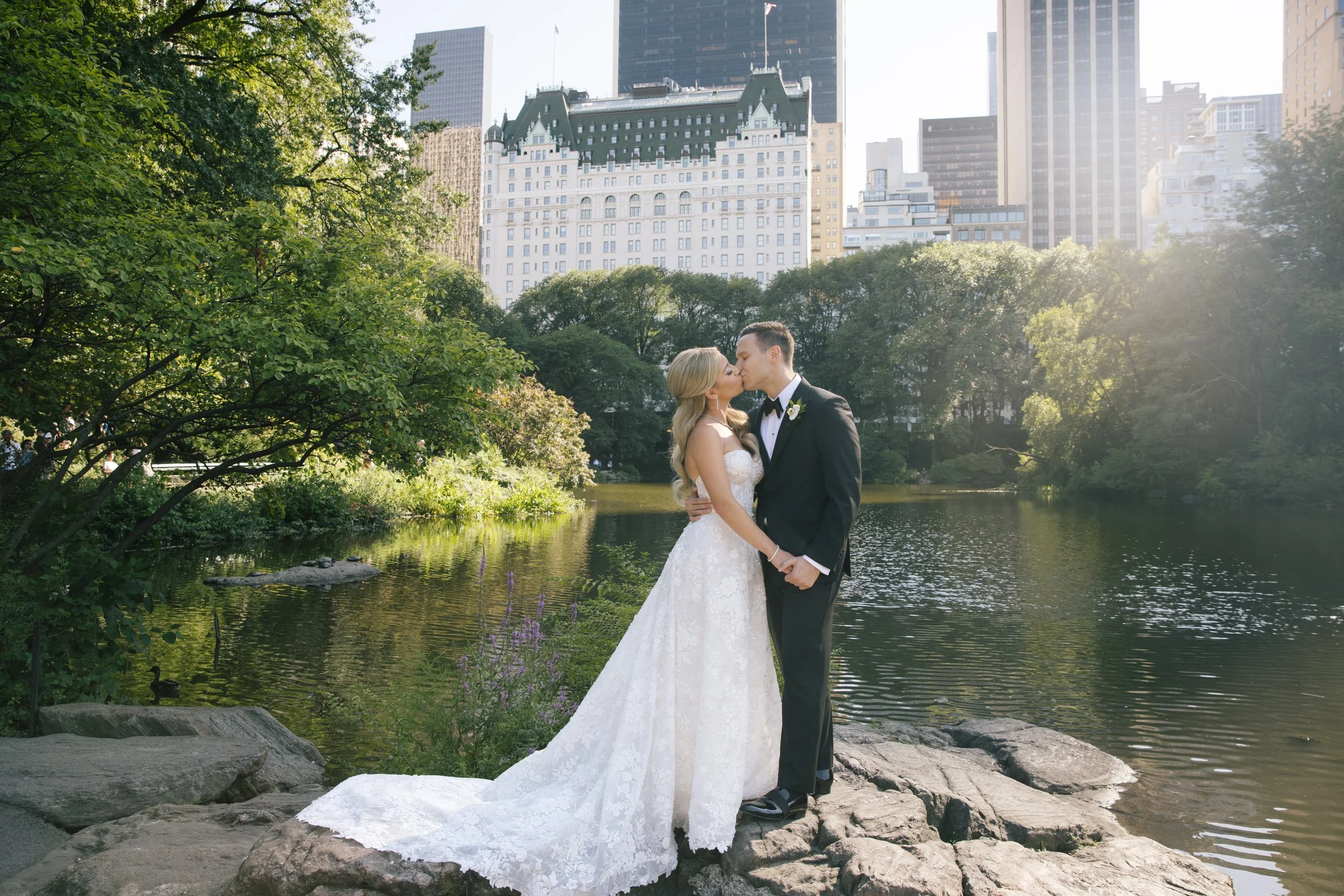 A bride and groom kiss by a pond in a park, with tall city buildings in the background.