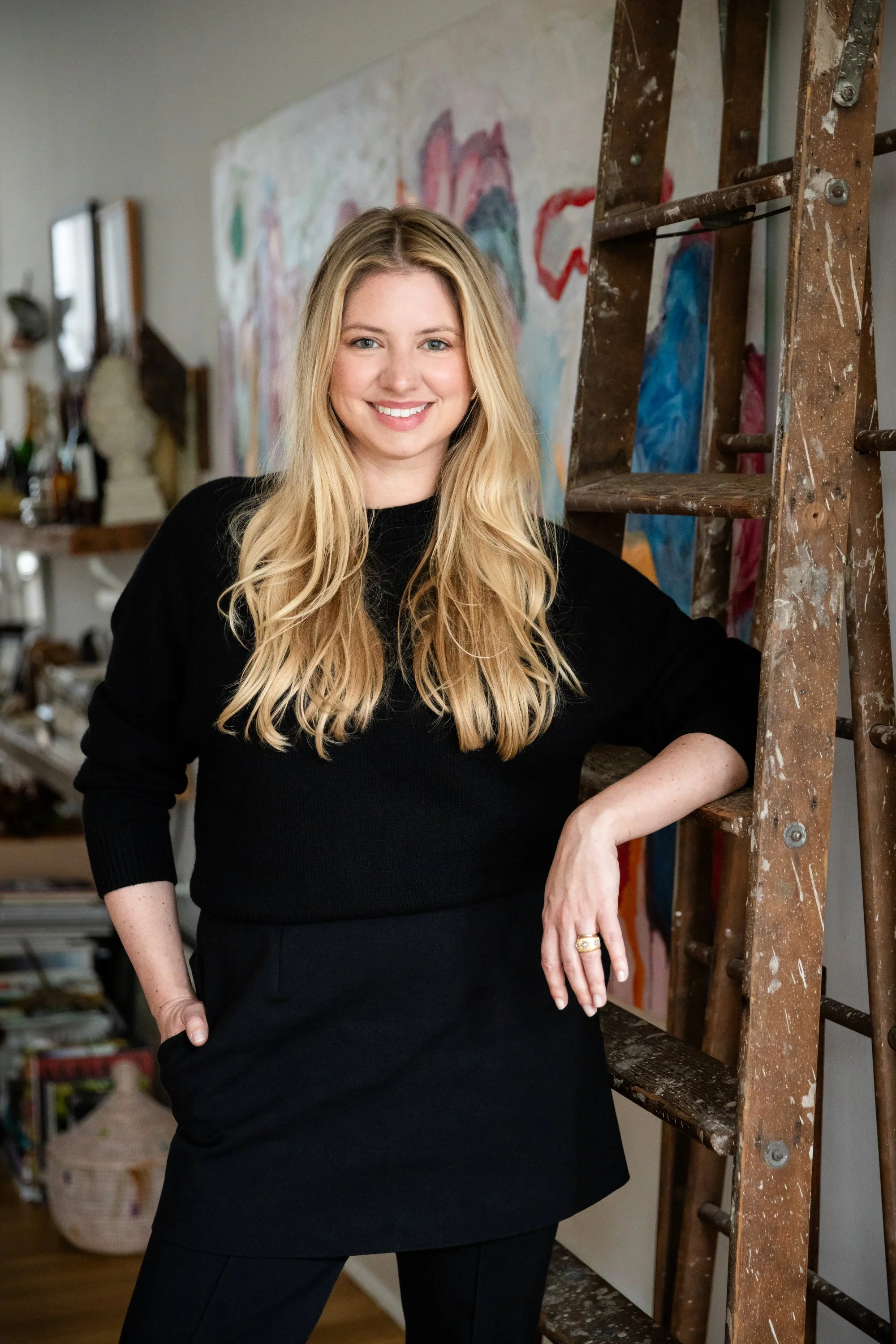 A woman with long blonde hair, wearing a black top and black pants, smiling and leaning against a wooden ladder, in an art studio with colorful paintings and sculptures in the background.