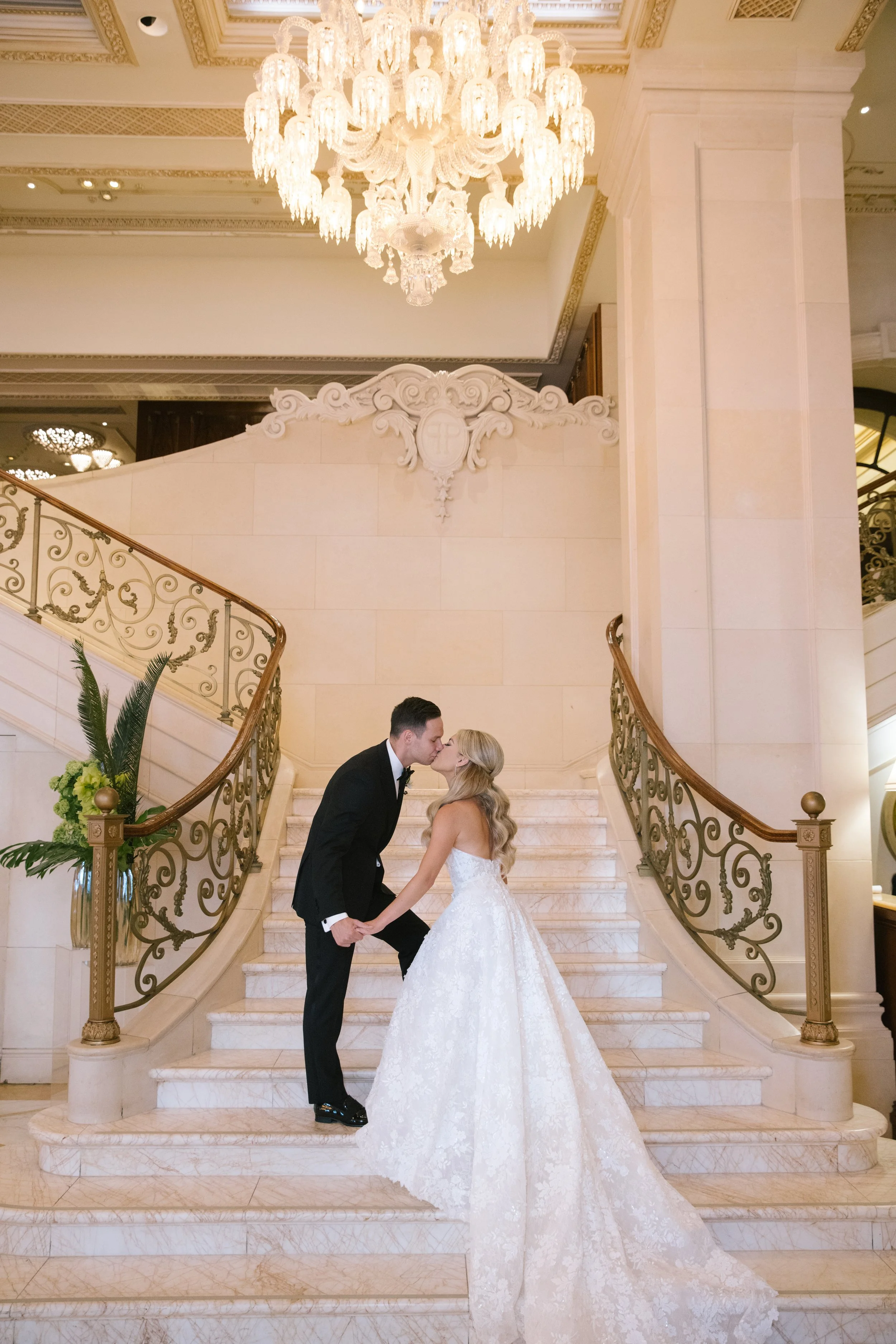 A newlywed couple dressed in wedding attire, holding hands and leaning in for a kiss on a grand staircase inside an elegant, ornate building with a large chandelier overhead.