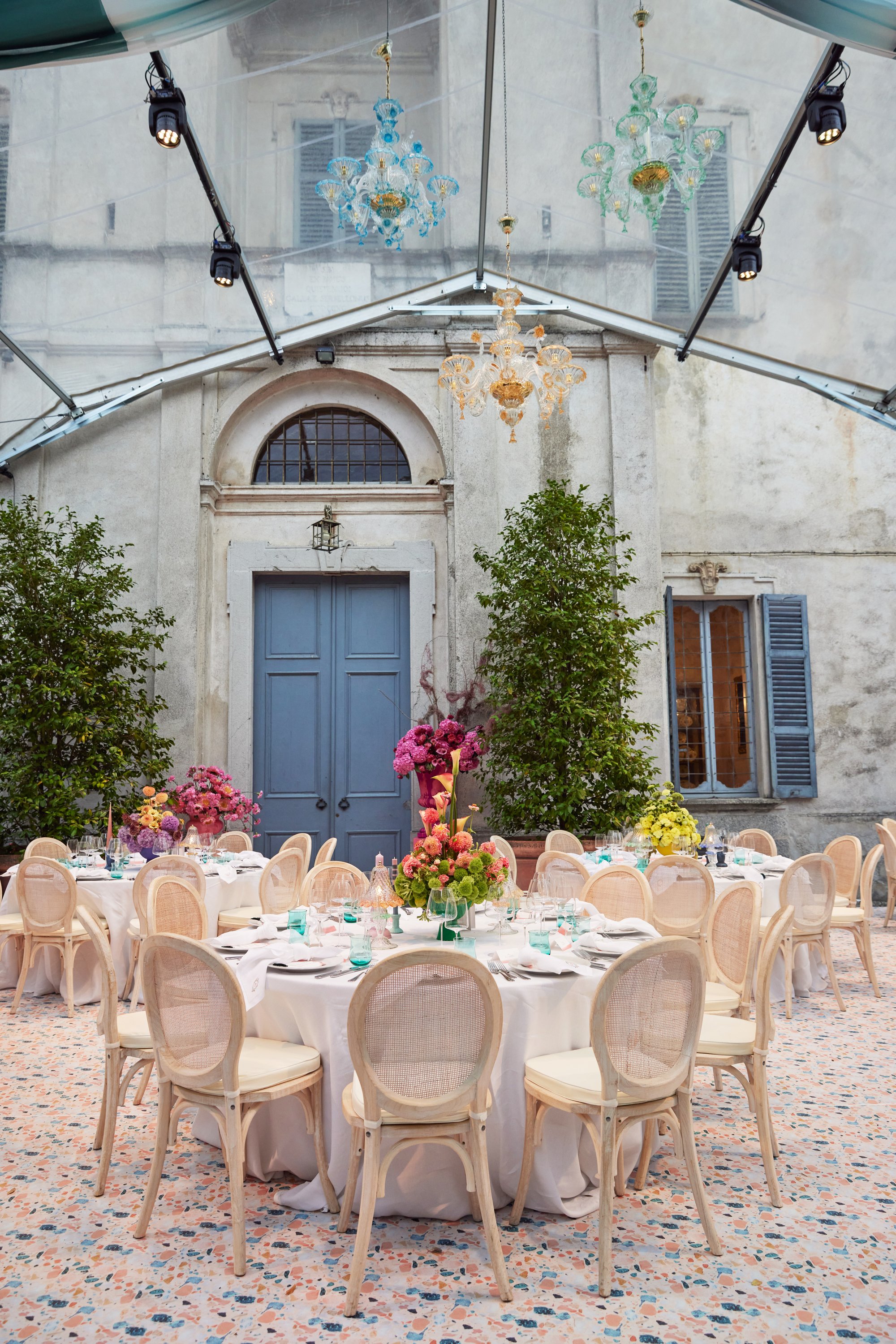 Elegant outdoor dining setup with round tables, white tablecloths, and beige chairs, decorated with colorful floral centerpieces under a transparent canopy. Old building facade with blue door, window shutters, and chandeliers hanging inside the canopy.