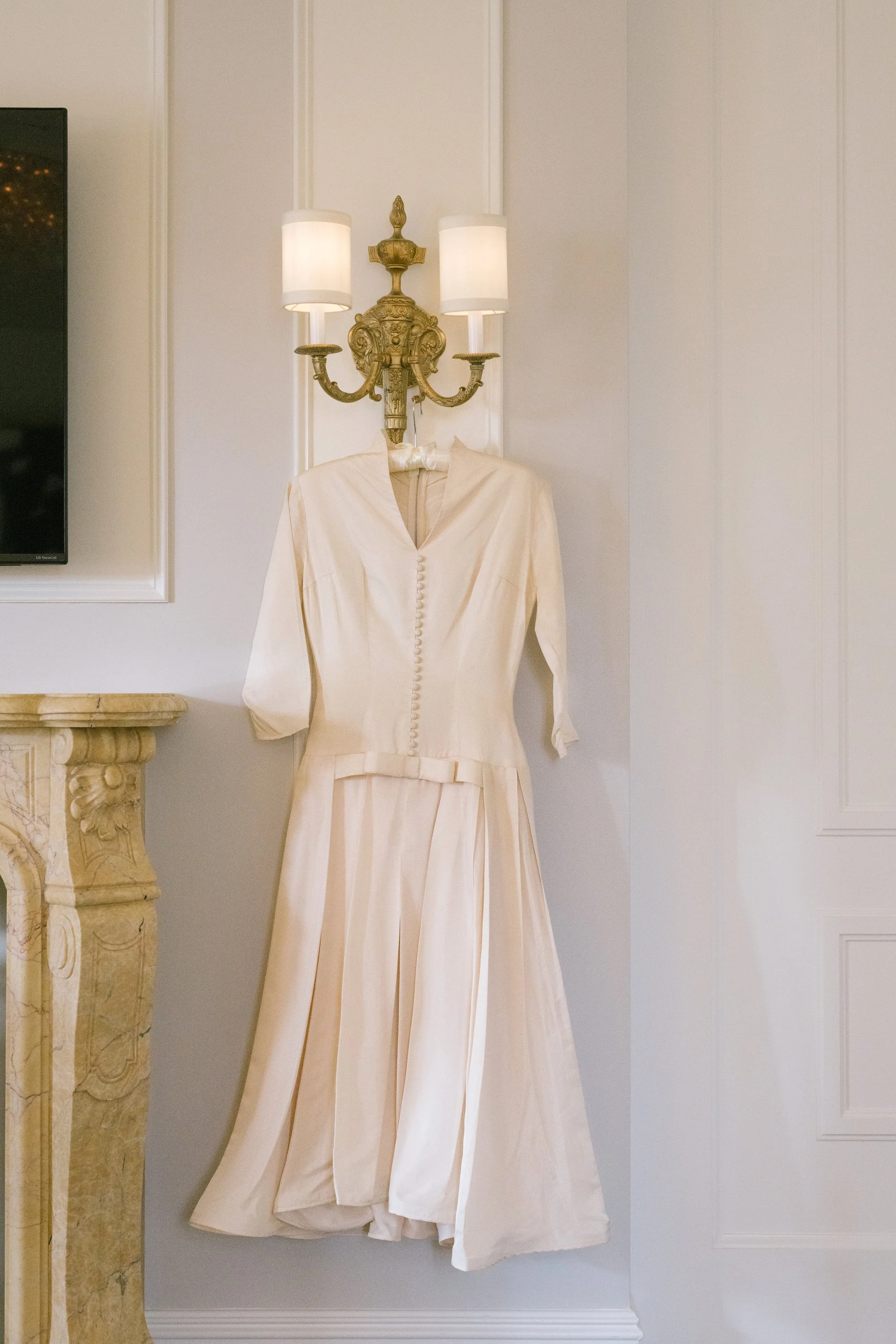 A vintage gold wall sconce with two white lampshades is mounted on a white wall. A cream-colored vintage dress with buttons down the front is hanging underneath the sconce. Part of a marble fireplace is visible on the left.
