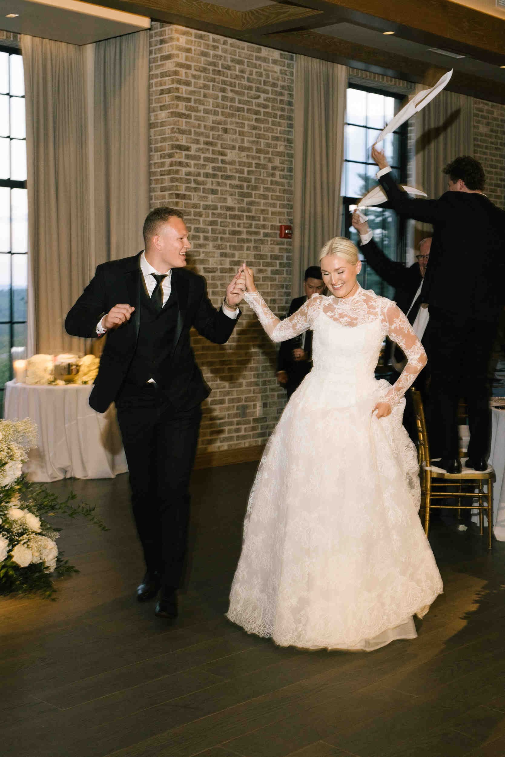 A bride and groom dancing at their wedding reception, smiling and holding hands, with a feathered decor element being held by a guest in the background.