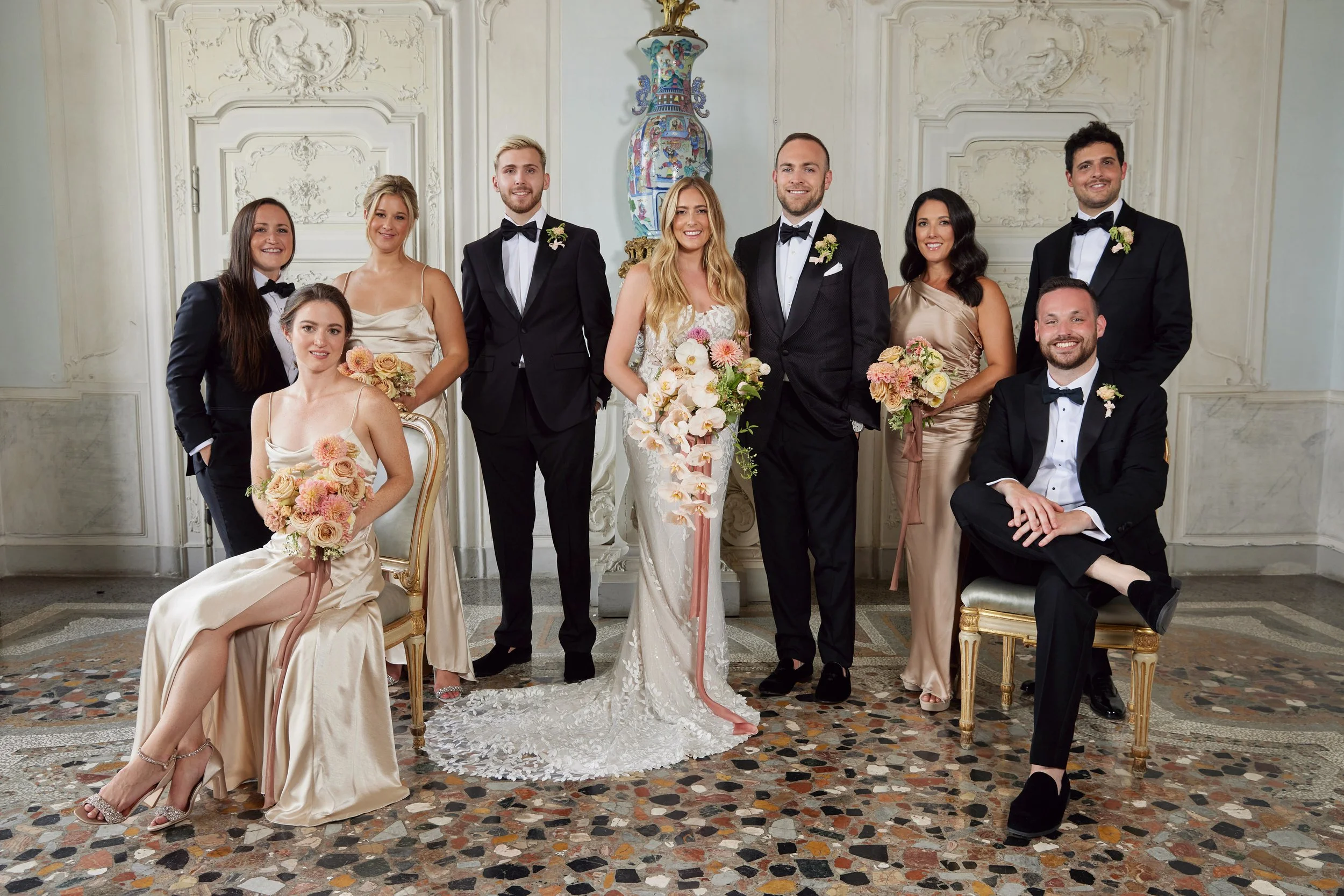 A group of ten people dressed in wedding attire posing in an ornate room with white walls and intricate moldings. The bride and groom are in the center, surrounded by bridesmaids and groomsmen.