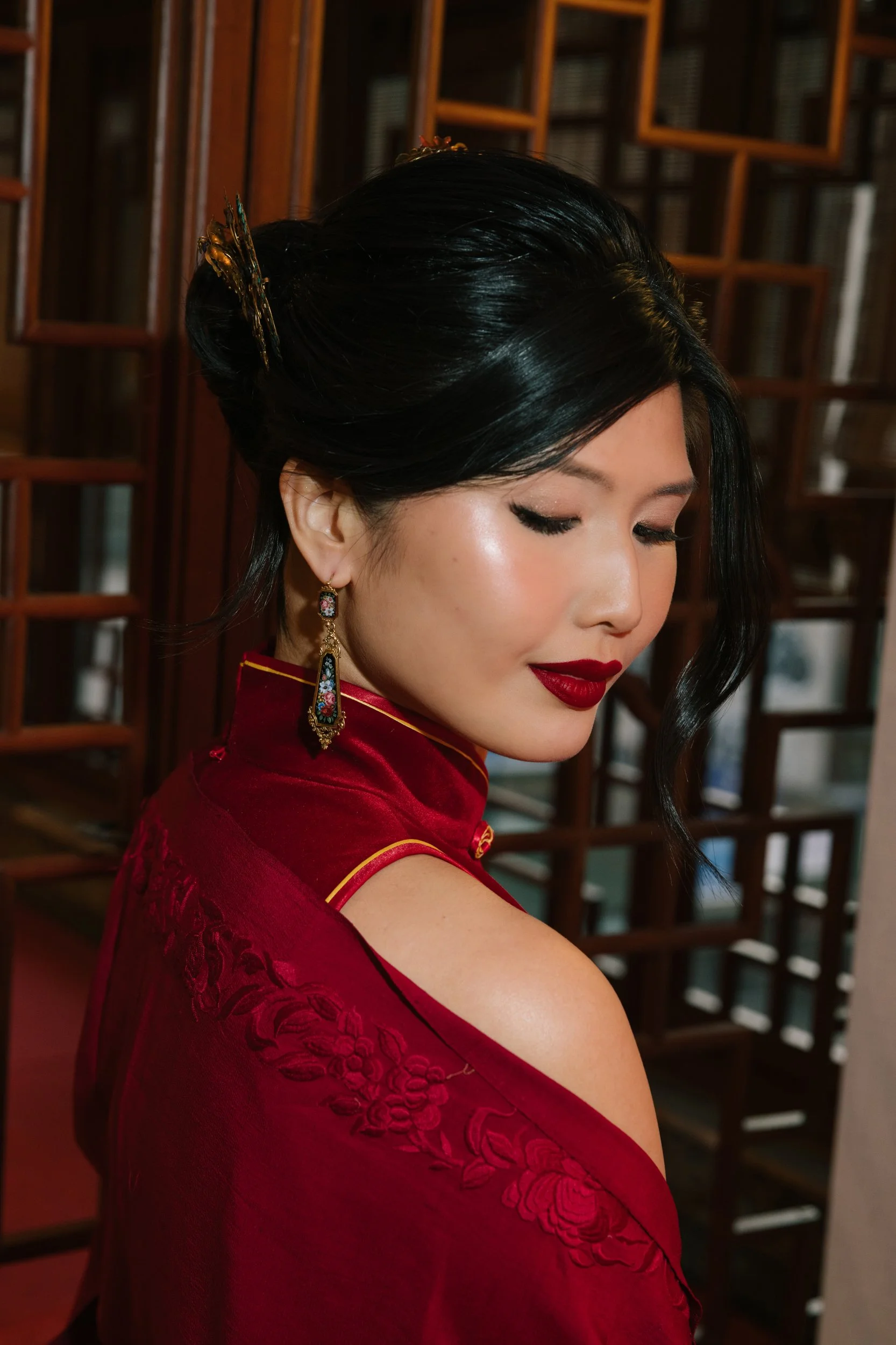 A woman with black hair styled in an elegant updo wearing traditional Chinese dress with intricate embroidery, red lipstick, and decorative earrings, standing in front of a wooden lattice background.