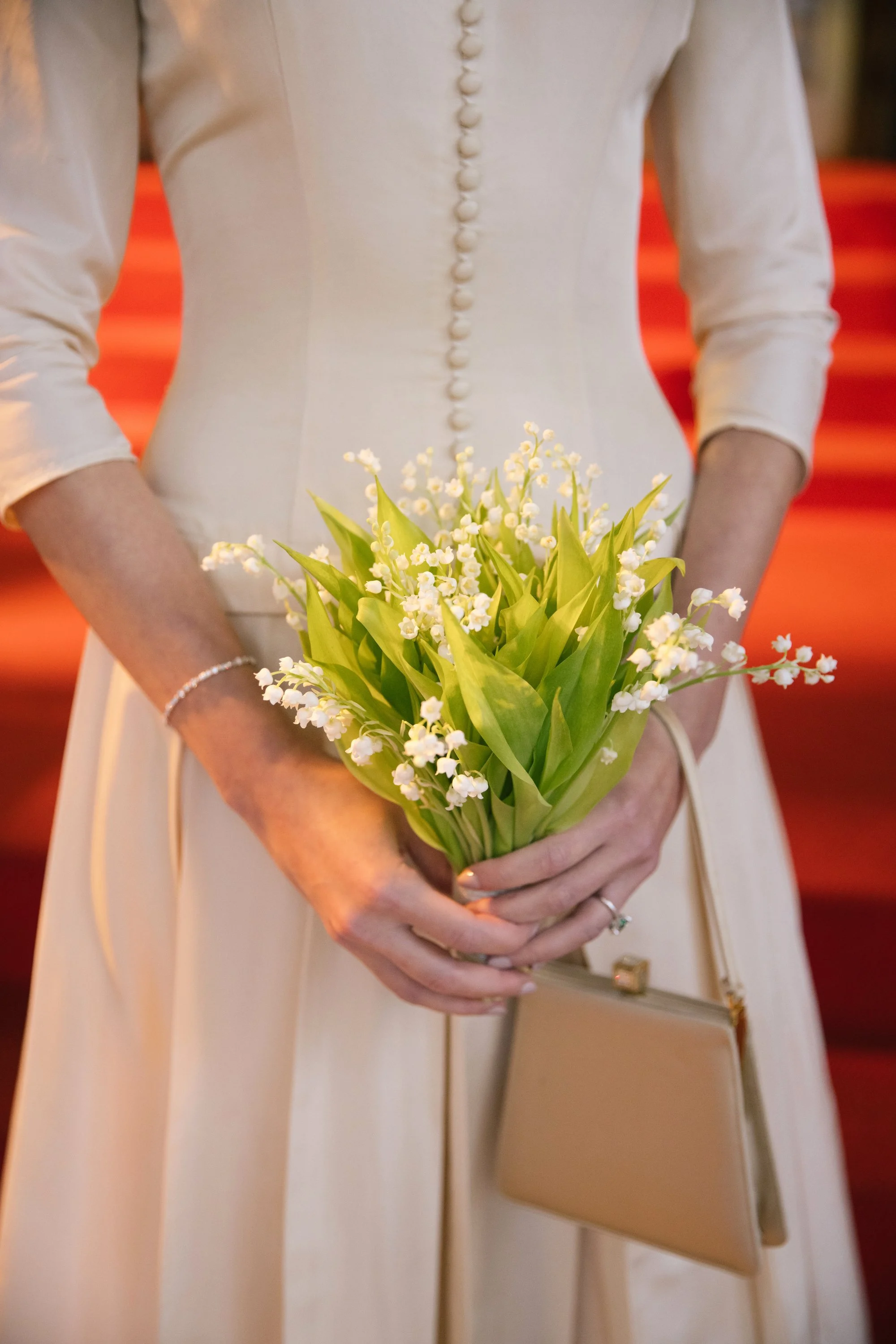 A woman in a cream-colored dress holding a bouquet of white lilies with green leaves, standing on red carpeted stairs.