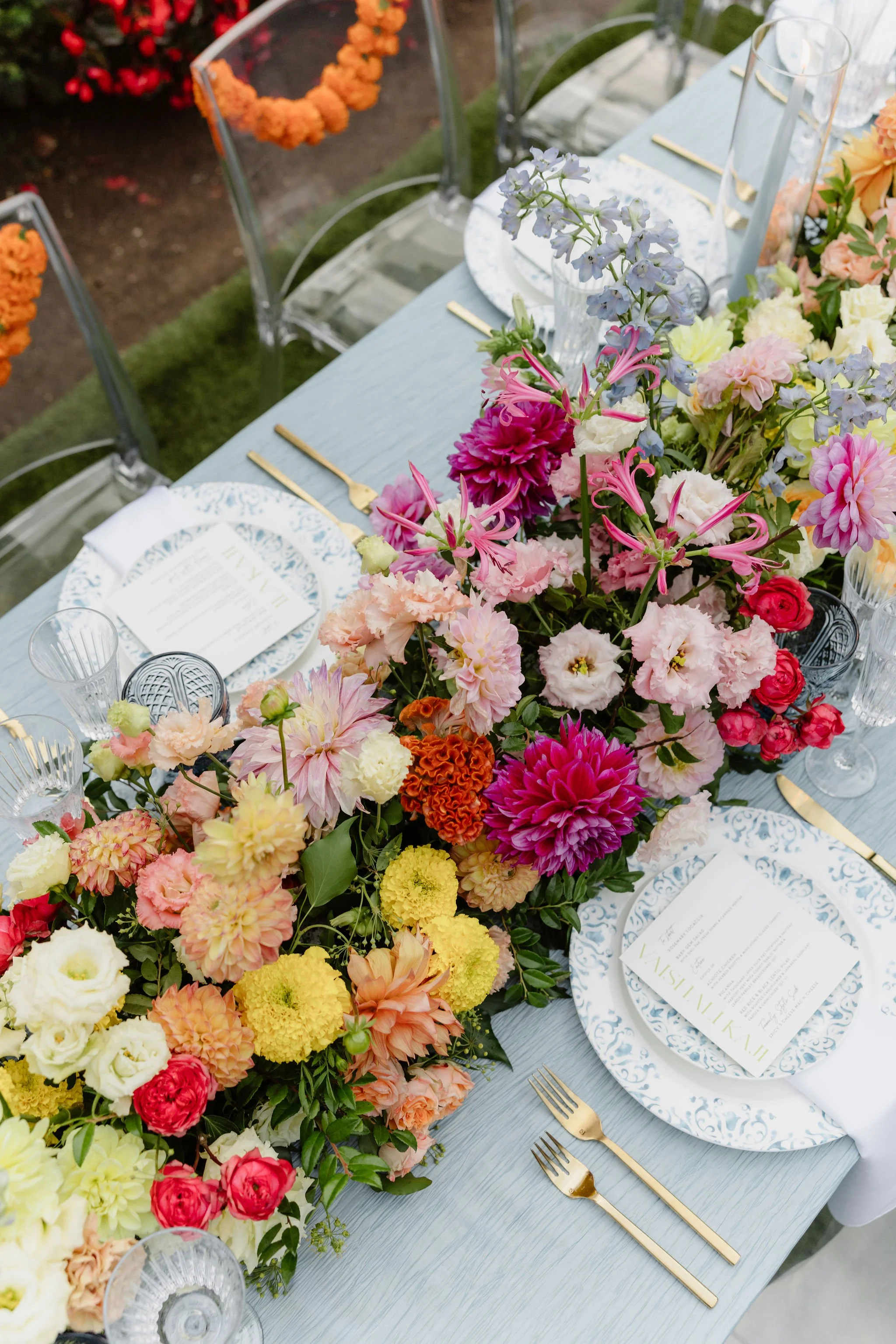 A beautifully decorated outdoor dining table with a large floral centerpiece, white and blue patterned plates, gold cutlery, and clear glassware.