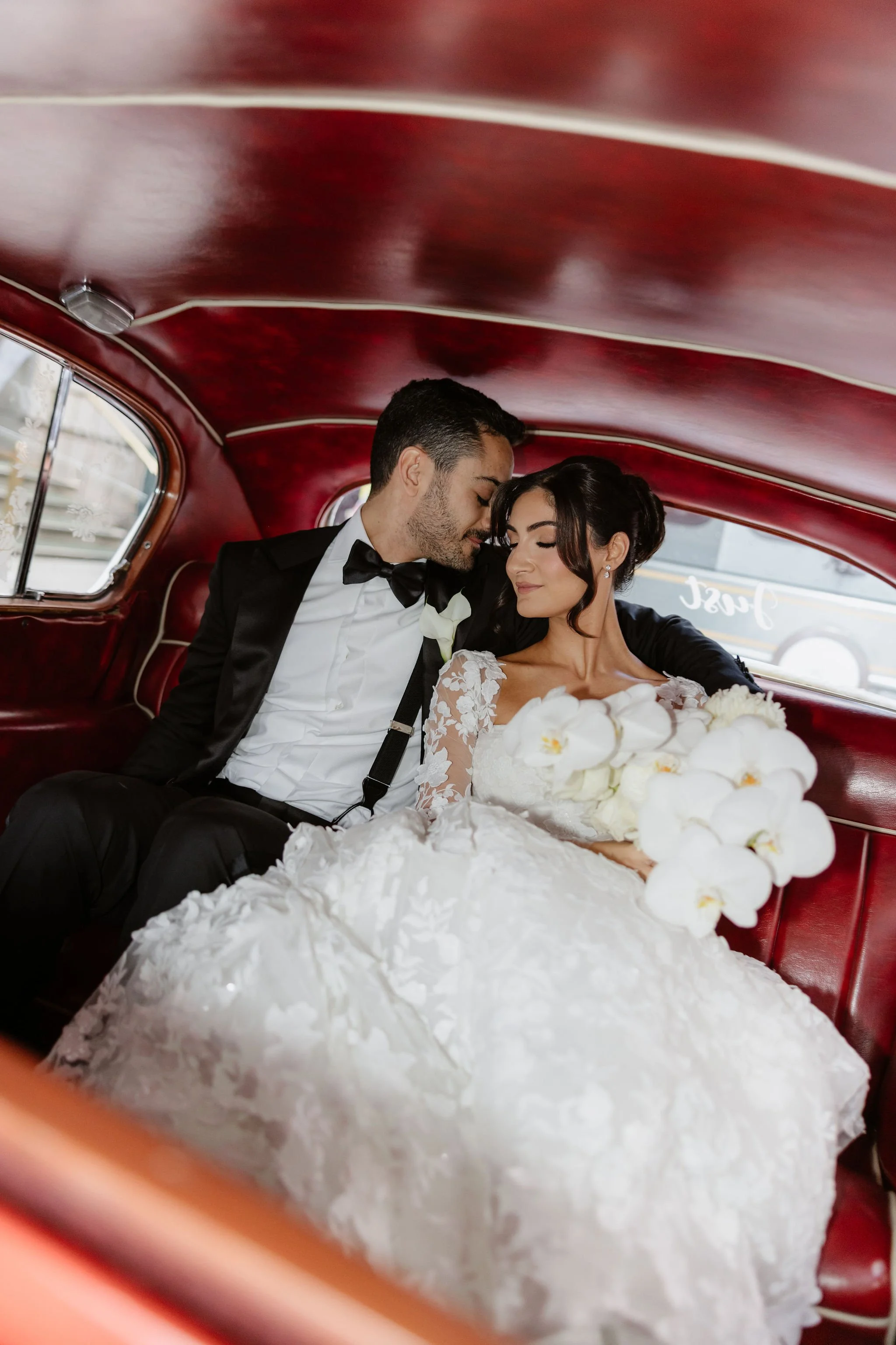 A newlywed couple in wedding attire sitting inside a vintage red car, looking content and close, with a bouquet of white flowers on the bride's lap.