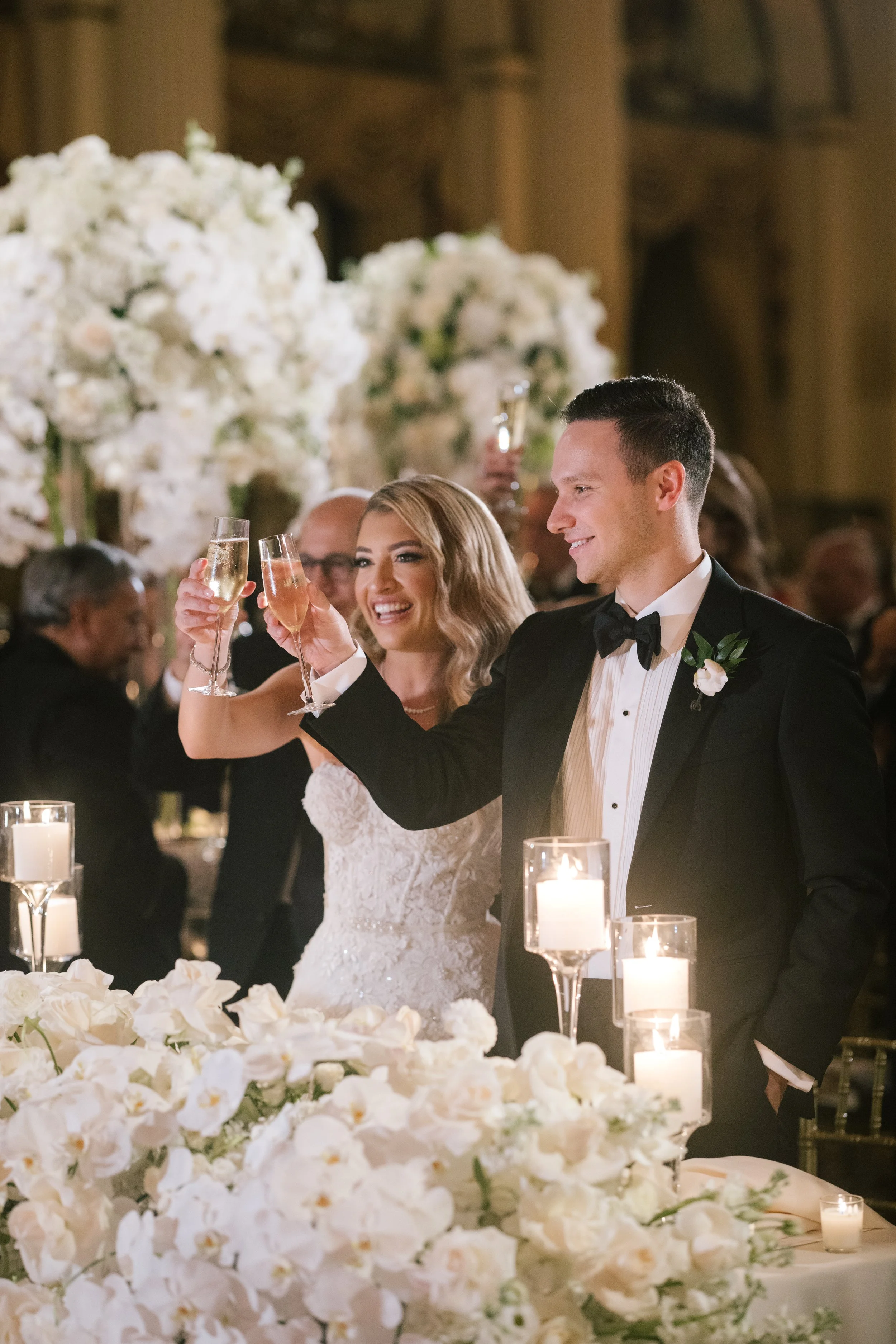 A newlywed couple raising glasses in a toast at their wedding reception, surrounded by white floral arrangements and candlelit decor.