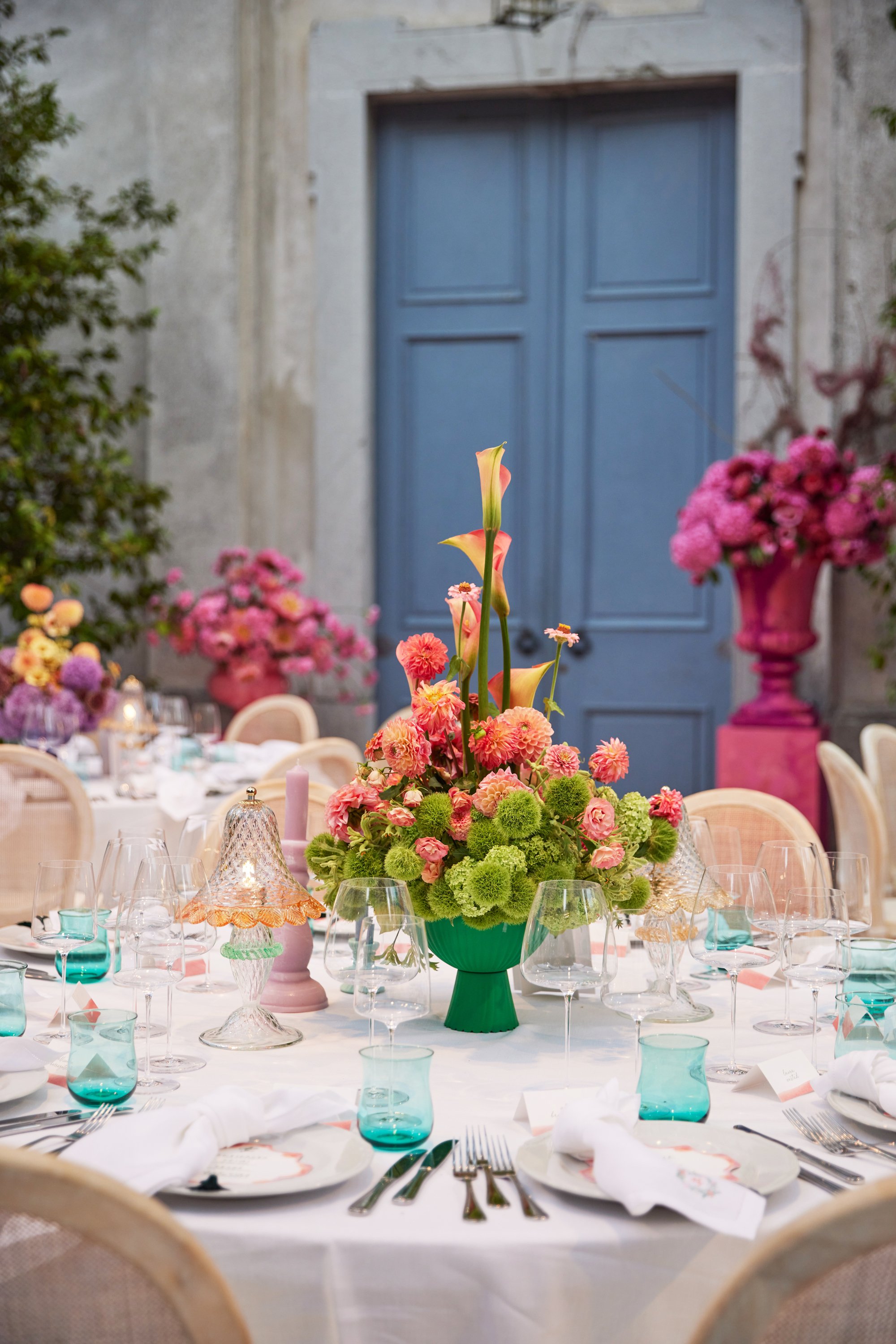 A round dining table decorated with a large floral centerpiece in a green vase, with pink, peach, and green flowers. The table is set with white napkins, plates, silverware, and teal glasses, in an elegant setting with a blue door in the background and pink and purple floral arrangements around the room.