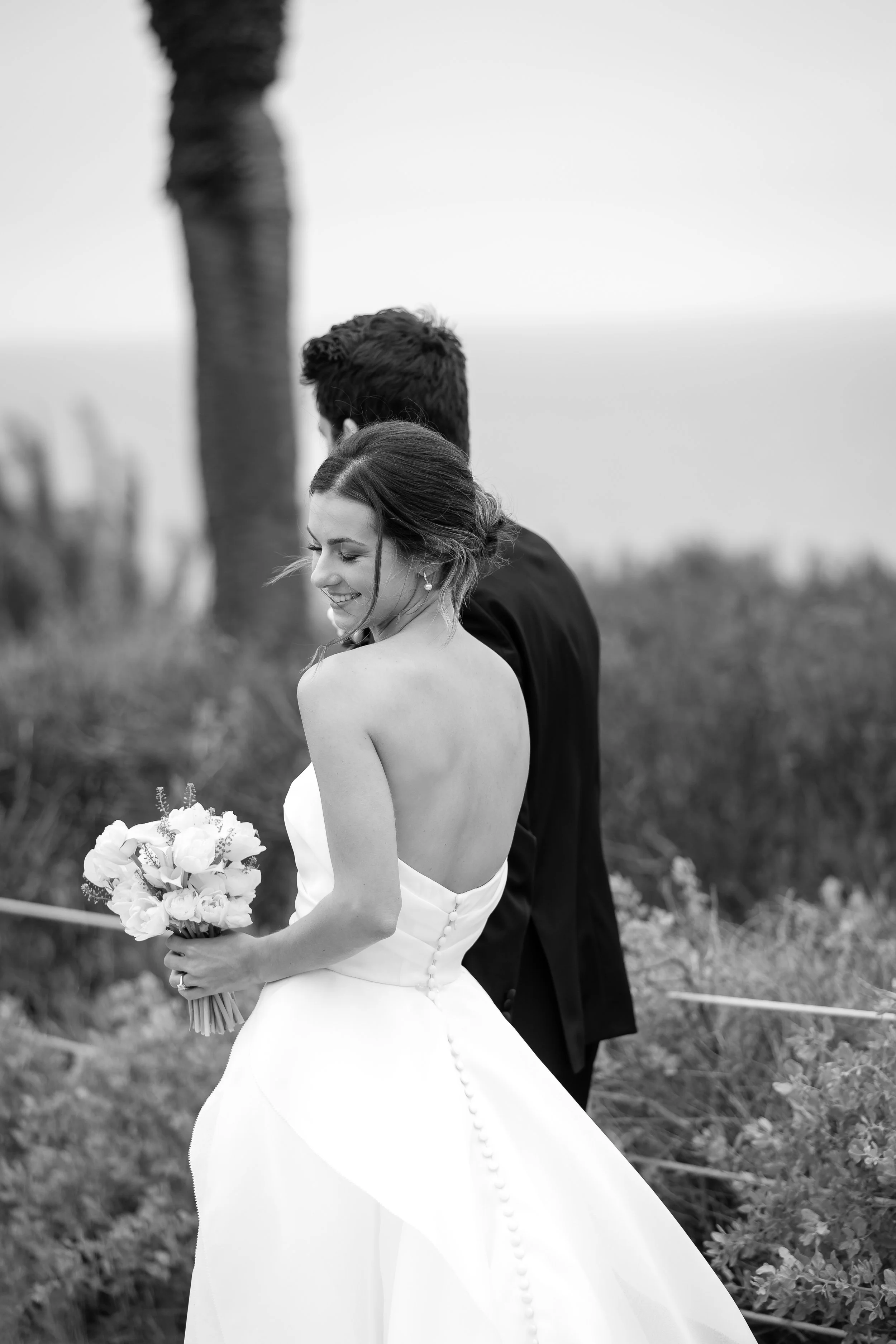 A black-and-white photo of a bride holding a bouquet of flowers, with her back partially exposed, and a groom standing behind her in an outdoor setting.
