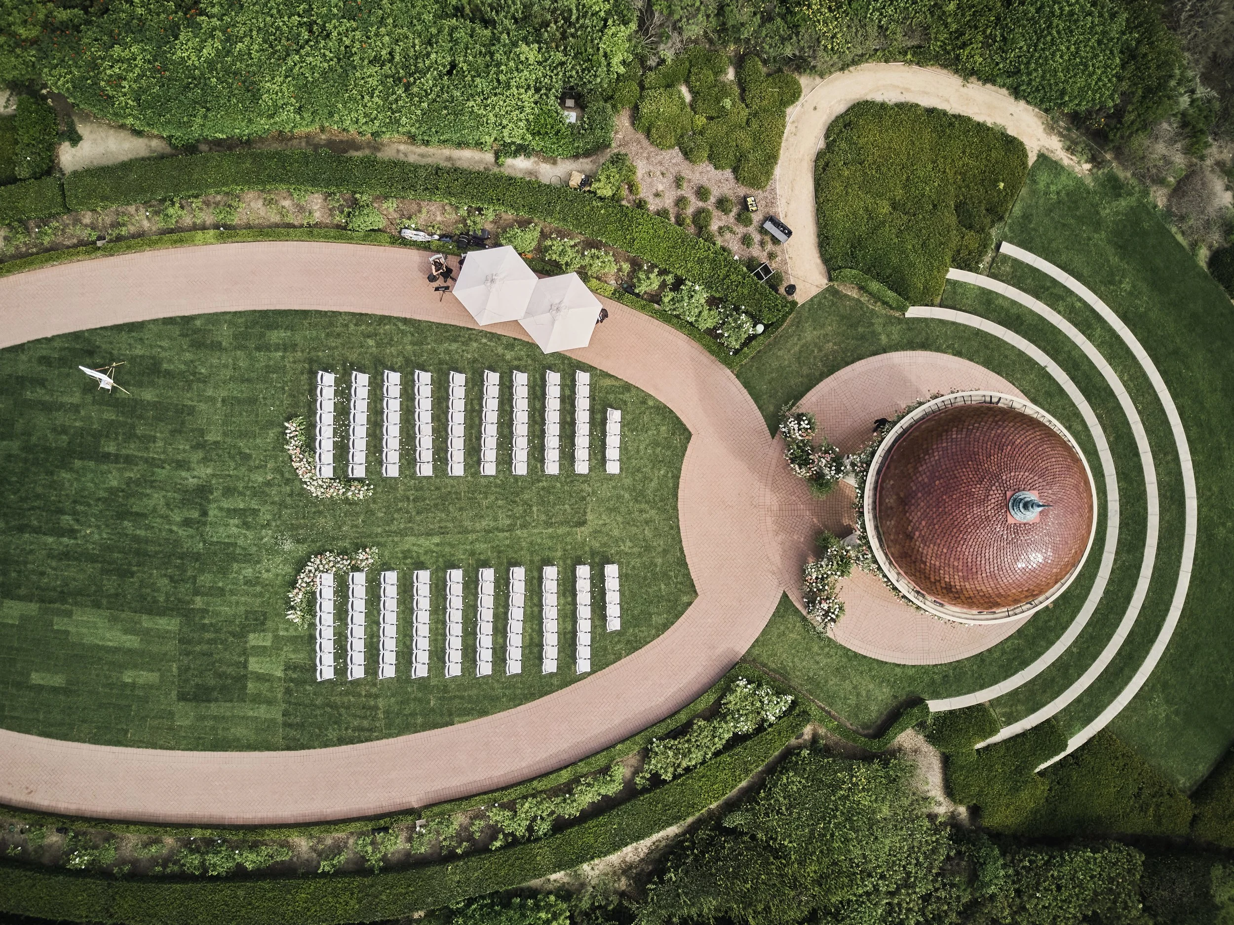 An aerial view of a park or garden with a wide brick pathway, white chairs arranged in rows, a small white tent, a curved pathway, and a domed brick structure surrounded by green trees and shrubs.
