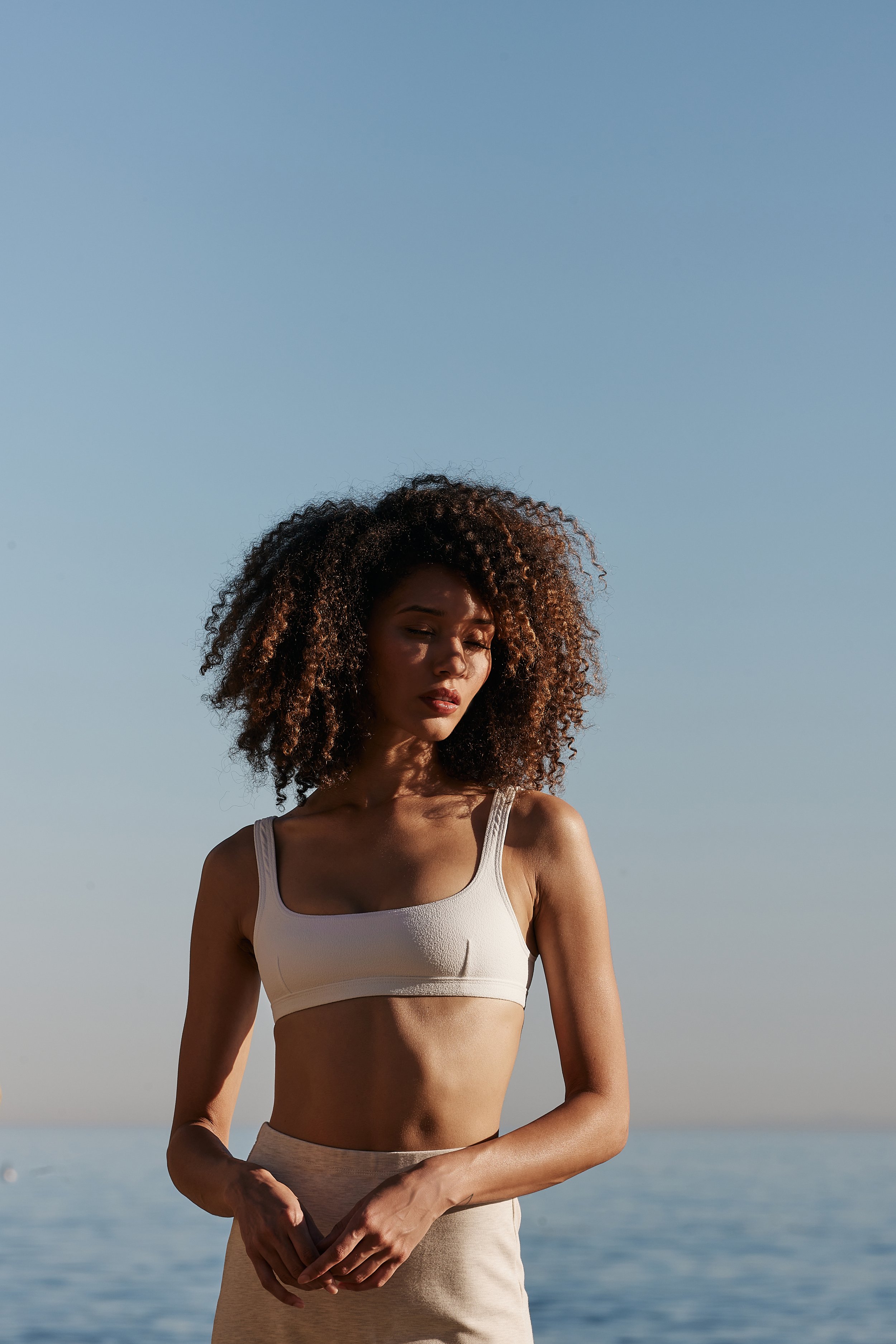 A woman with curly hair wearing a peach sports bra and matching shorts stands outdoors near water under a clear blue sky.