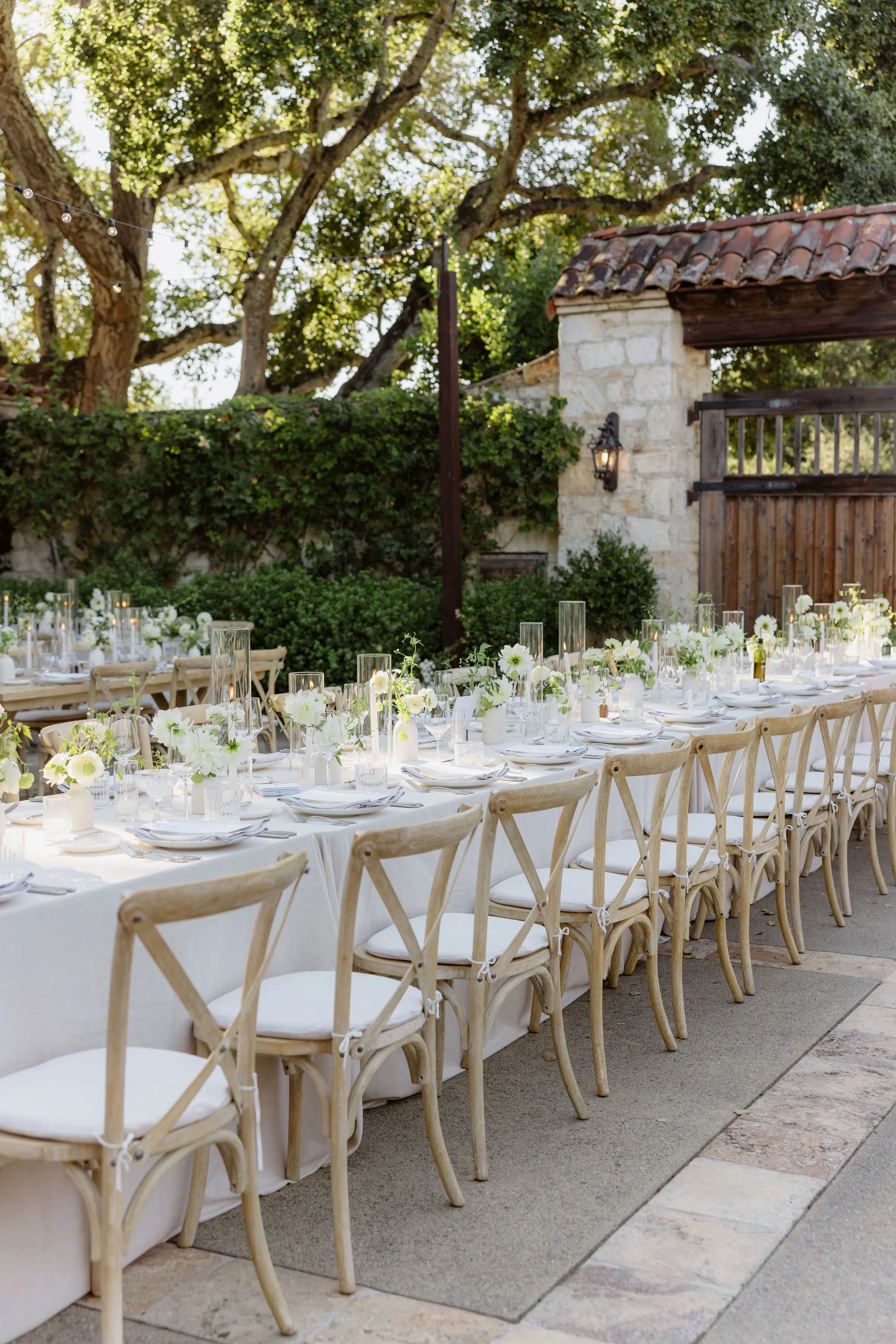 Outdoor dining setup with long table covered in white tablecloth, decorated with white flowers and candles, surrounded by wooden chairs under large trees with string lights and a stone wall and wooden gate in the background.