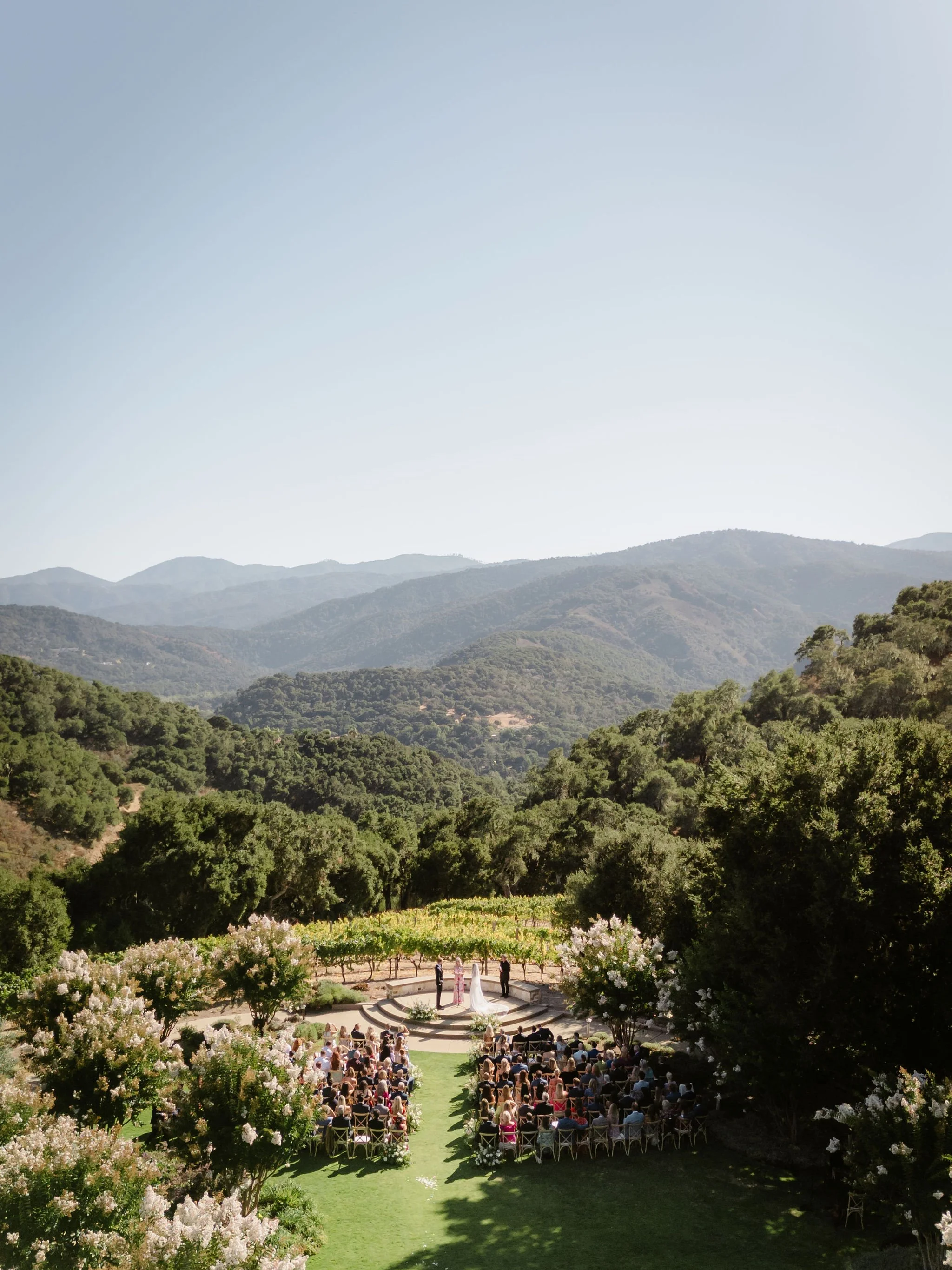 Wedding ceremony taking place outdoors in a scenic mountainous area with a ceremony site surrounded by blossoming trees, guests seated in rows, and mountains in the background.