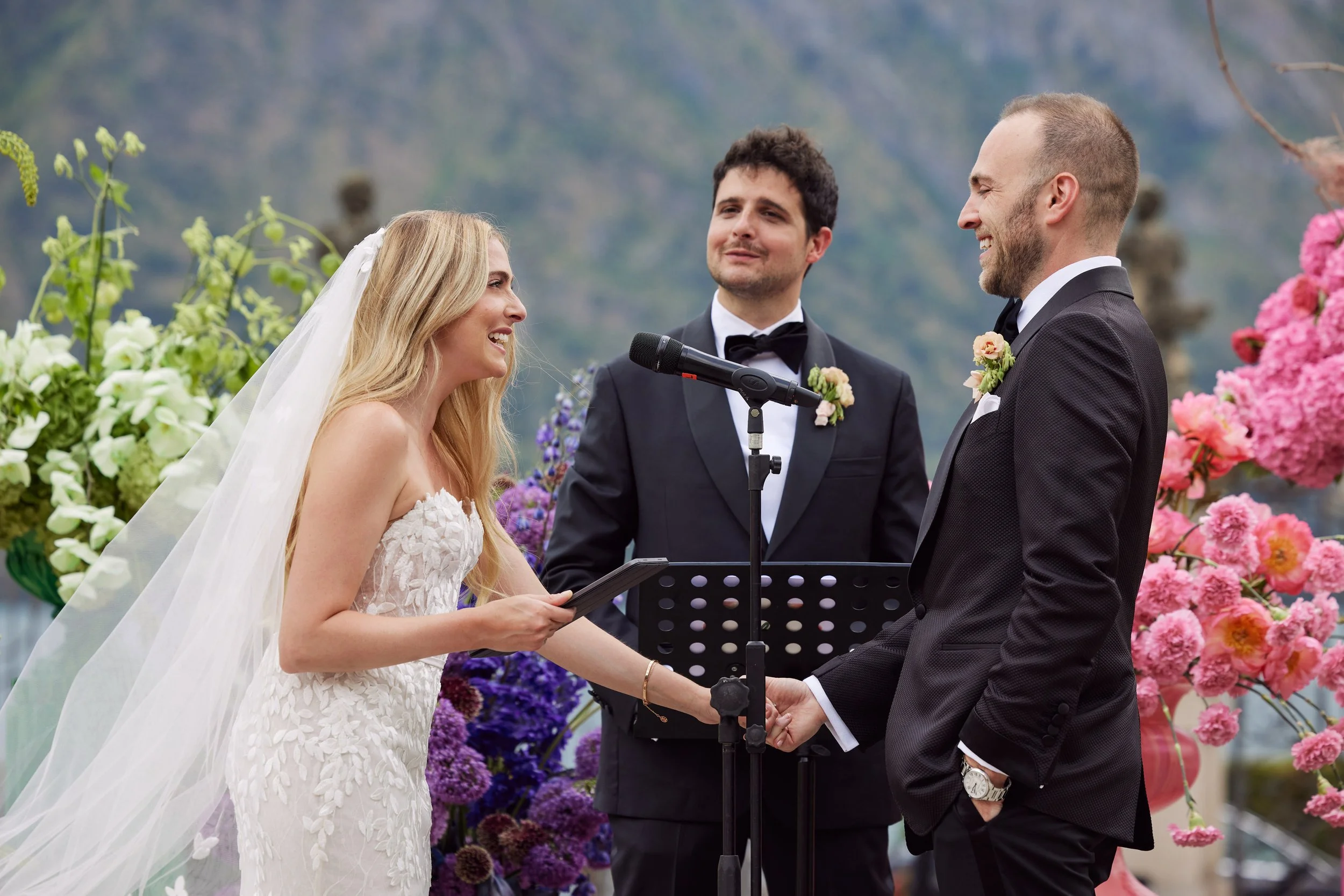 A bride and groom exchange vows during a wedding ceremony outdoors, with a officiant standing behind them. The bride wears a white wedding gown and veil, and the groom wears a black tuxedo. They are holding hands and smiling, surrounded by pink and p