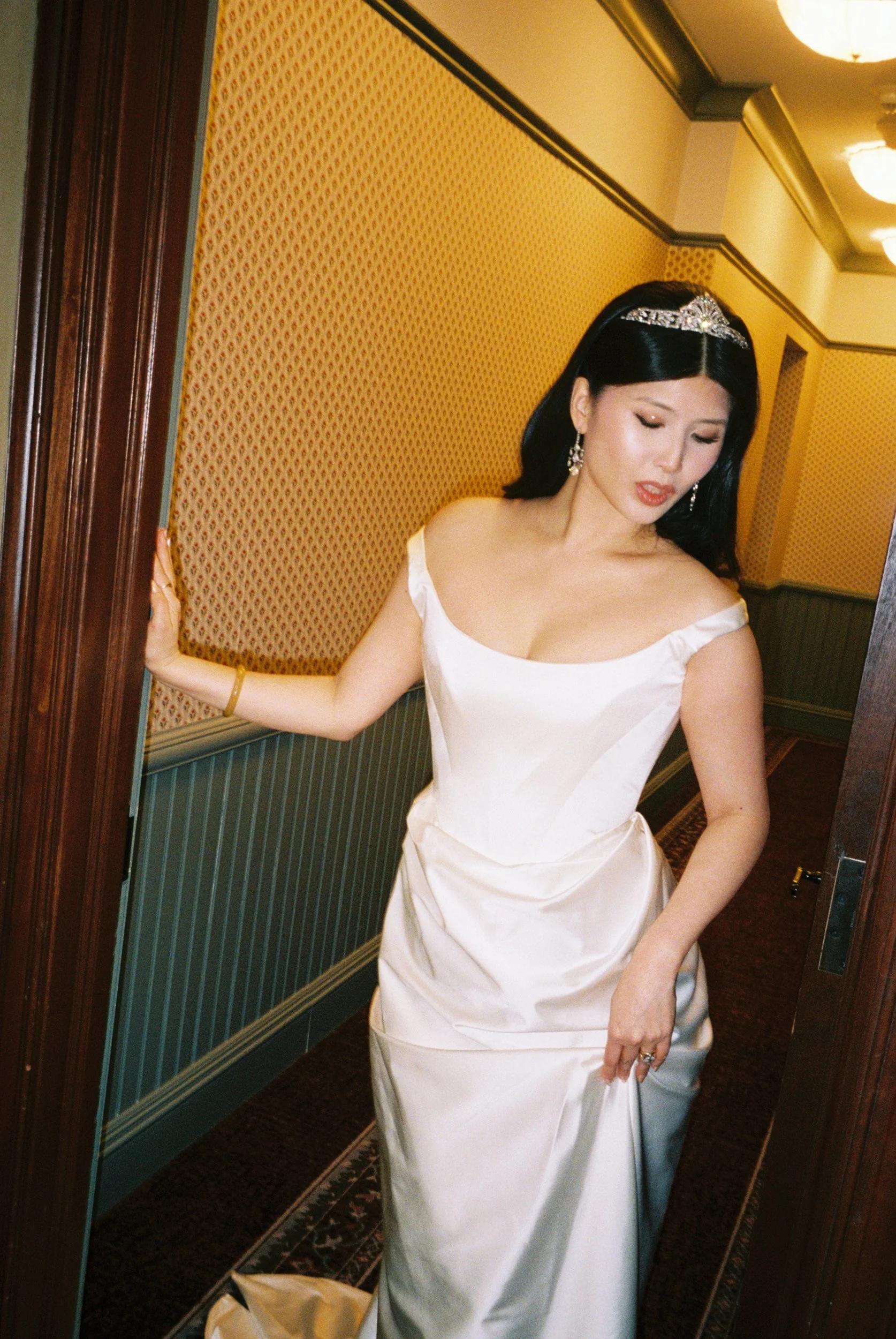 A woman in a white satin wedding dress with off-the-shoulder straps, wearing earrings and a tiara, standing in a hallway with yellow patterned wallpaper and dark wood trim, holding onto a door frame.
