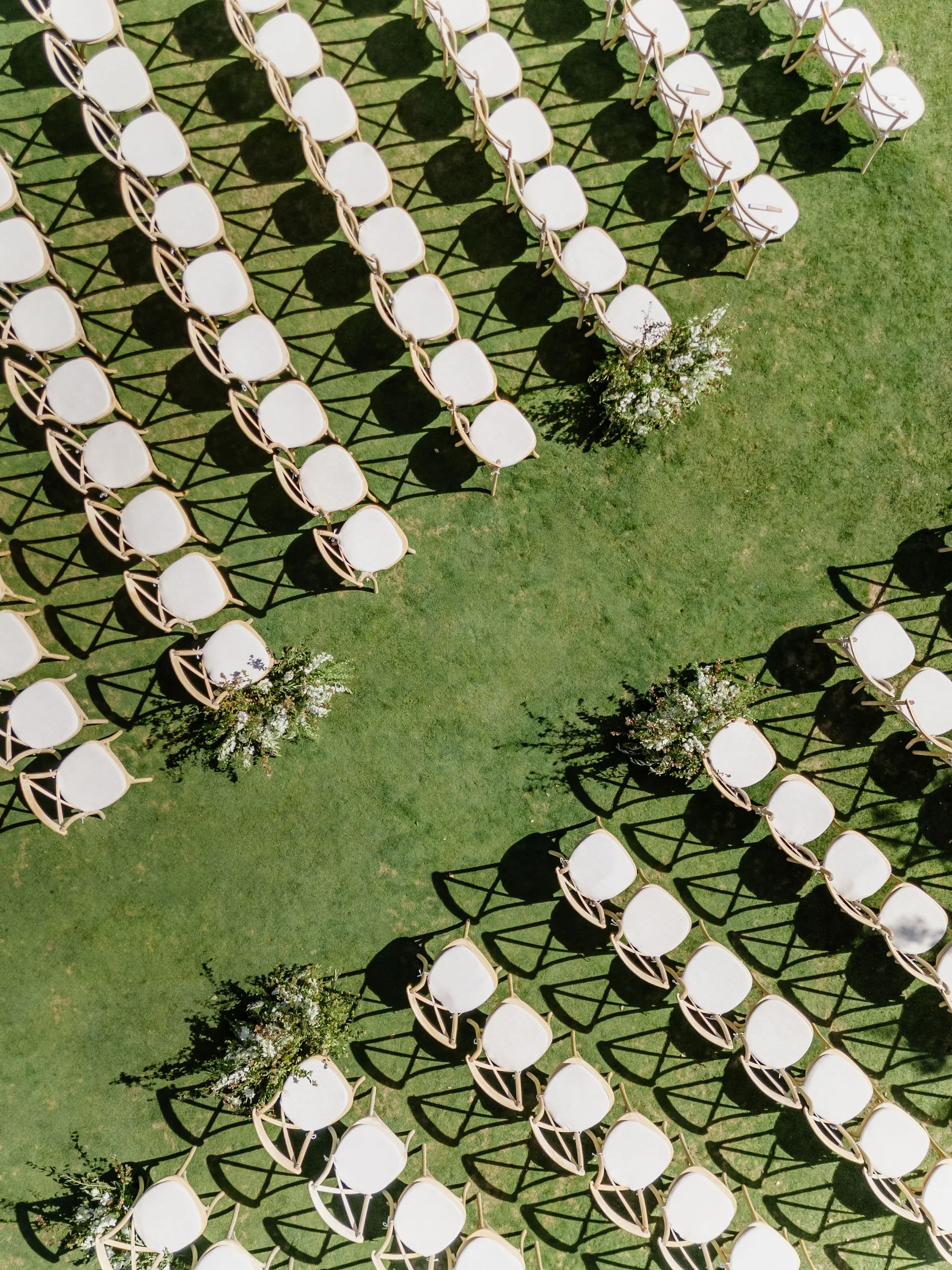 Empty white chairs arranged outdoors on green grass with small floral arrangements placed among the chairs and casting shadows in a pattern.