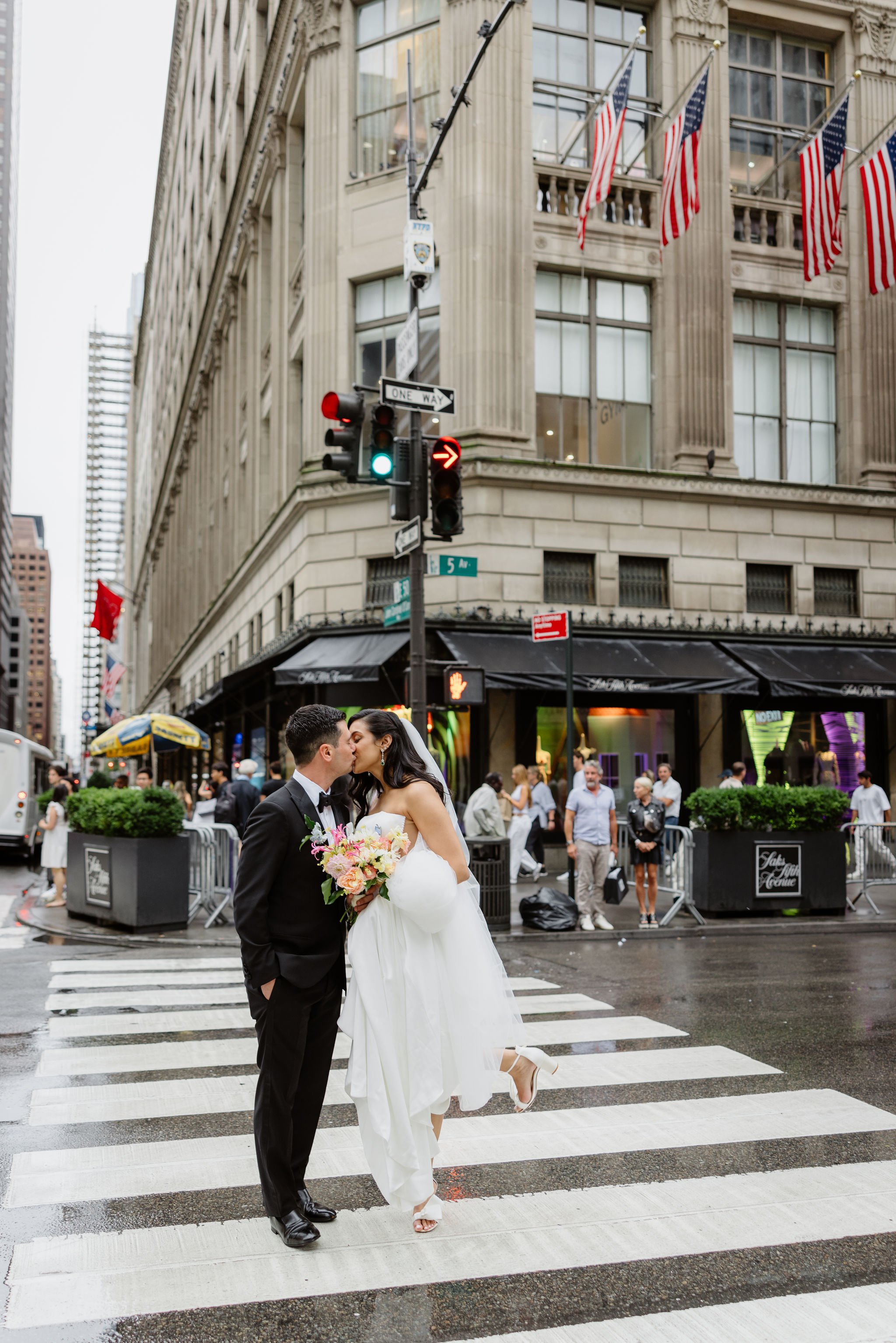 A newlywed couple sharing a kiss on a city street crossing, with the bride holding a bouquet of flowers and lifting one foot as she kisses the groom.