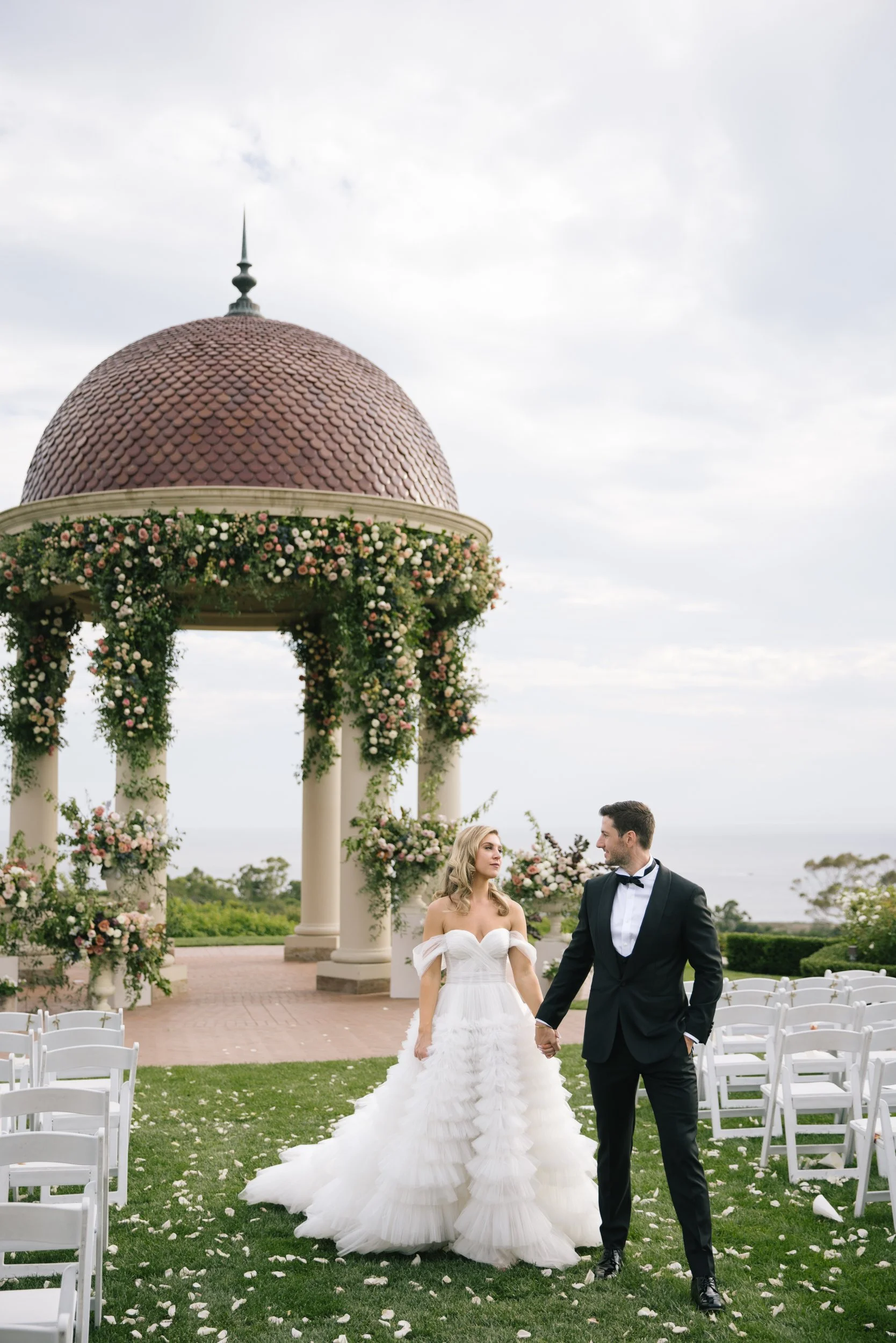 A bride and groom walk hand in hand through an outdoor wedding ceremony area decorated with white chairs and floral arrangements, with a decorative pavilion and cloudy sky in the background.