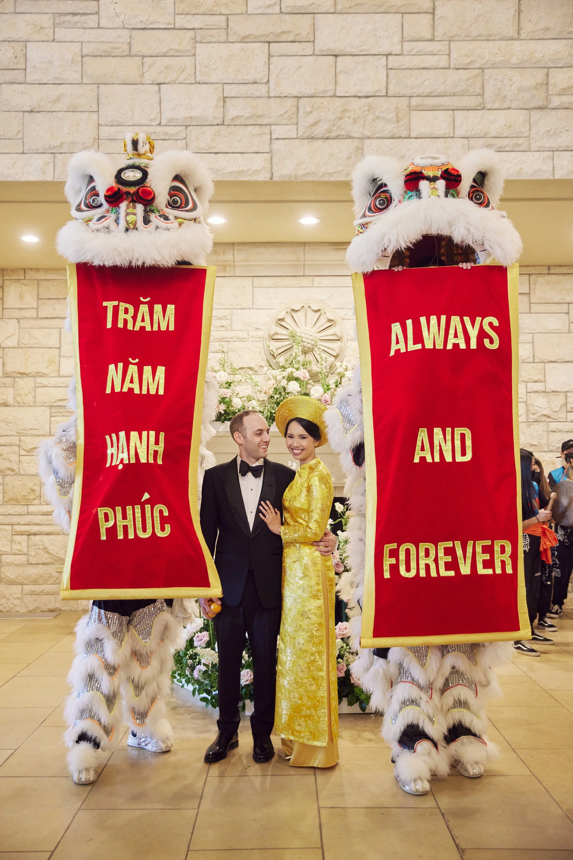 A couple dressed in formal attire standing between two traditional lion dance costumes with banners that read "Trăm Năm Hạnh Phúc" and "Always and Forever" at an indoor celebration.