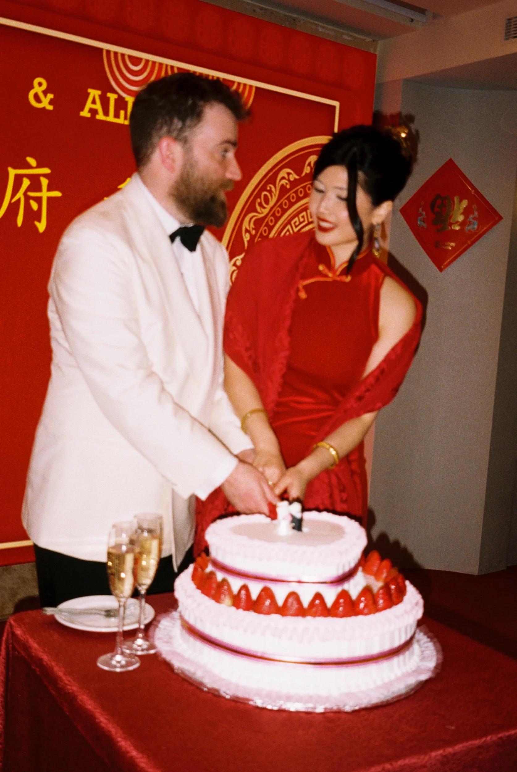 A couple dressed in formal attire cutting a large, decorated cake at a celebration. The woman is wearing a red dress with traditional Chinese design elements, and the man is wearing a white tuxedo with a black bow tie. There are two champagne glasses